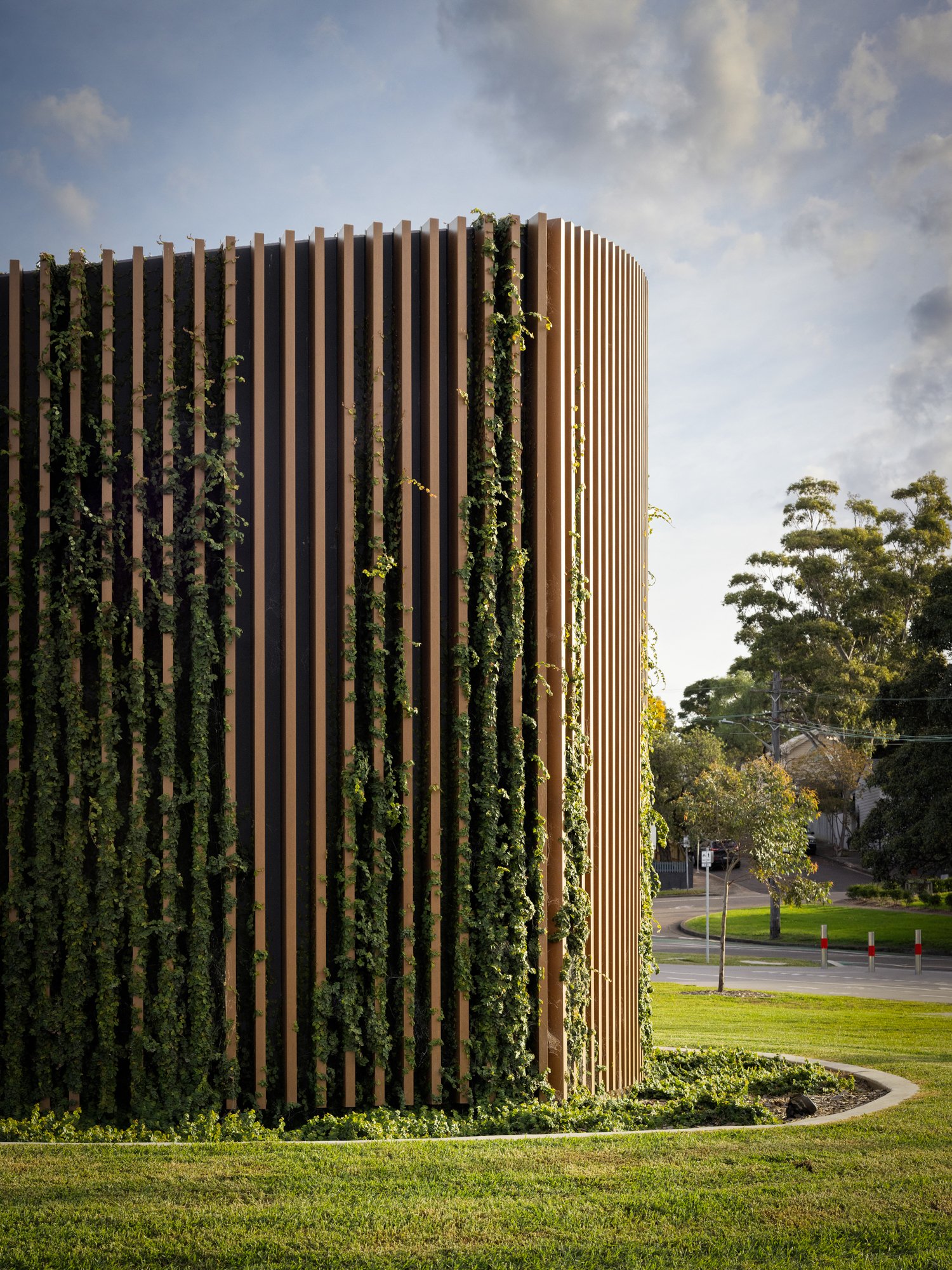 A modern building with vertical wooden slats and green vines growing on it, surrounded by a well-maintained lawn and trees in the background, under a partly cloudy sky. Designed by Colin Polwarth Studio Photo by Ruth Gold