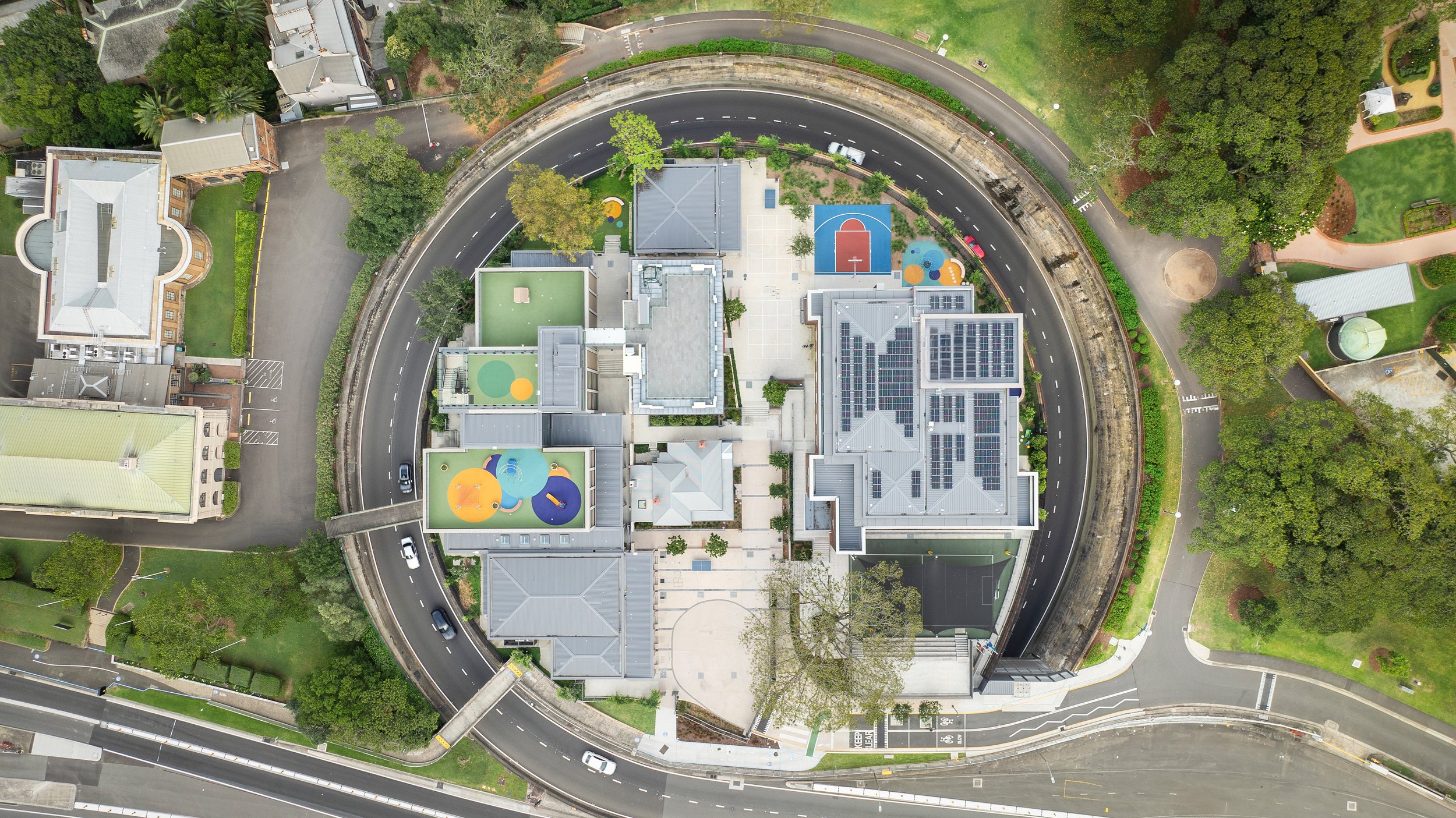 An aerial view of a school building with solar panels on the roof, surrounded by a circular road and parking lot, with playgrounds and green spaces nearby. Designed by fjcStudio Photo by Ruth Gold