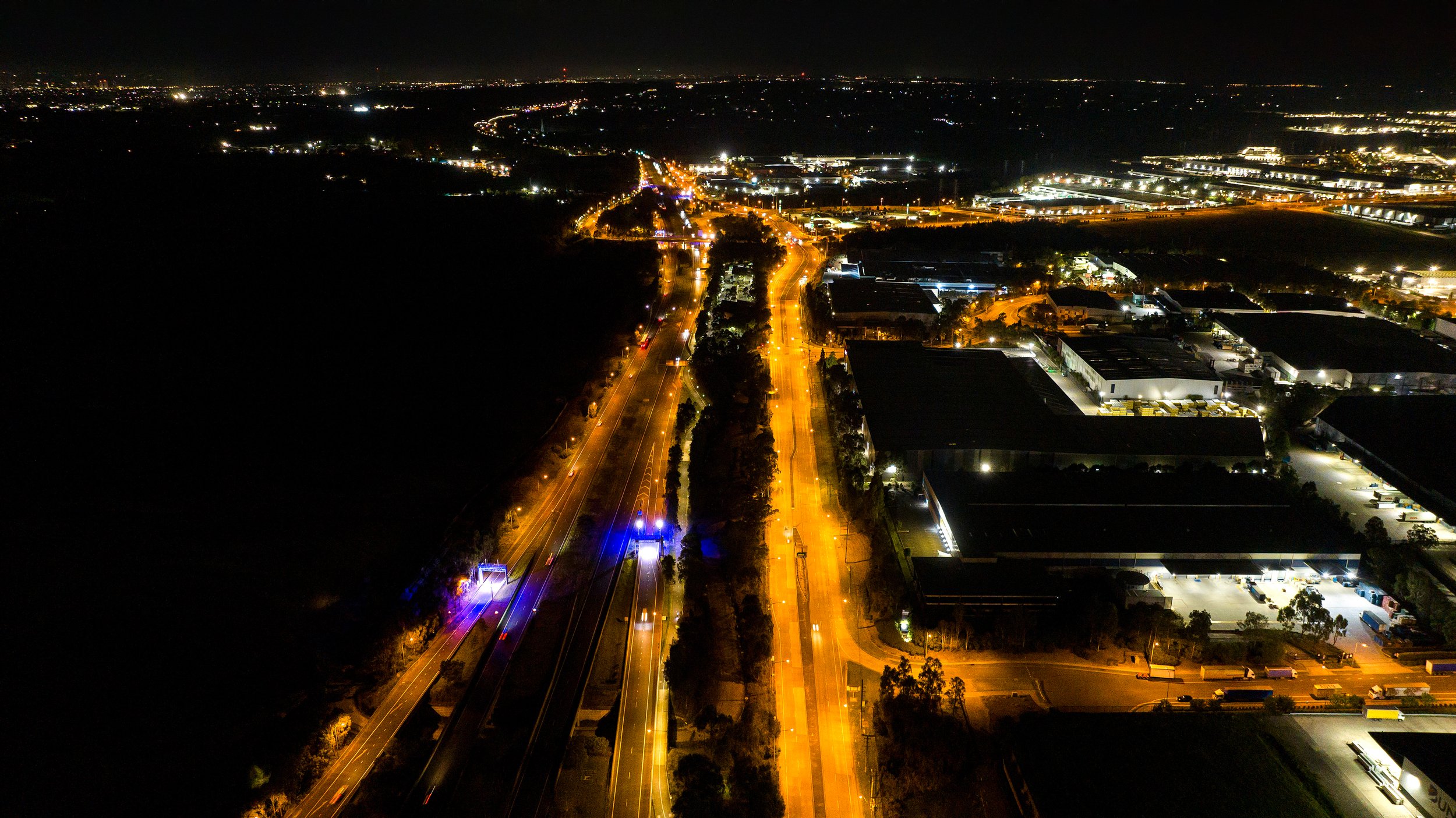 Night aerial view of a highway with streetlights and vehicles, surrounded by industrial and commercial buildings, with a dark landscape on the left and a cityscape with distant lights on the horizon. Designed by Colin Polwarth Studio Photo by Ruth Go