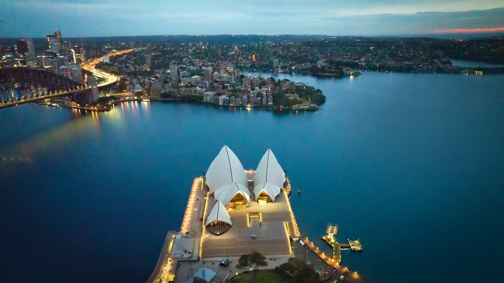 An aerial view of the Sydney Opera House illuminated at dusk, situated on the edge of Sydney Harbour with city buildings and bridges visible in the background. Opera House Photo by Ruth Gold