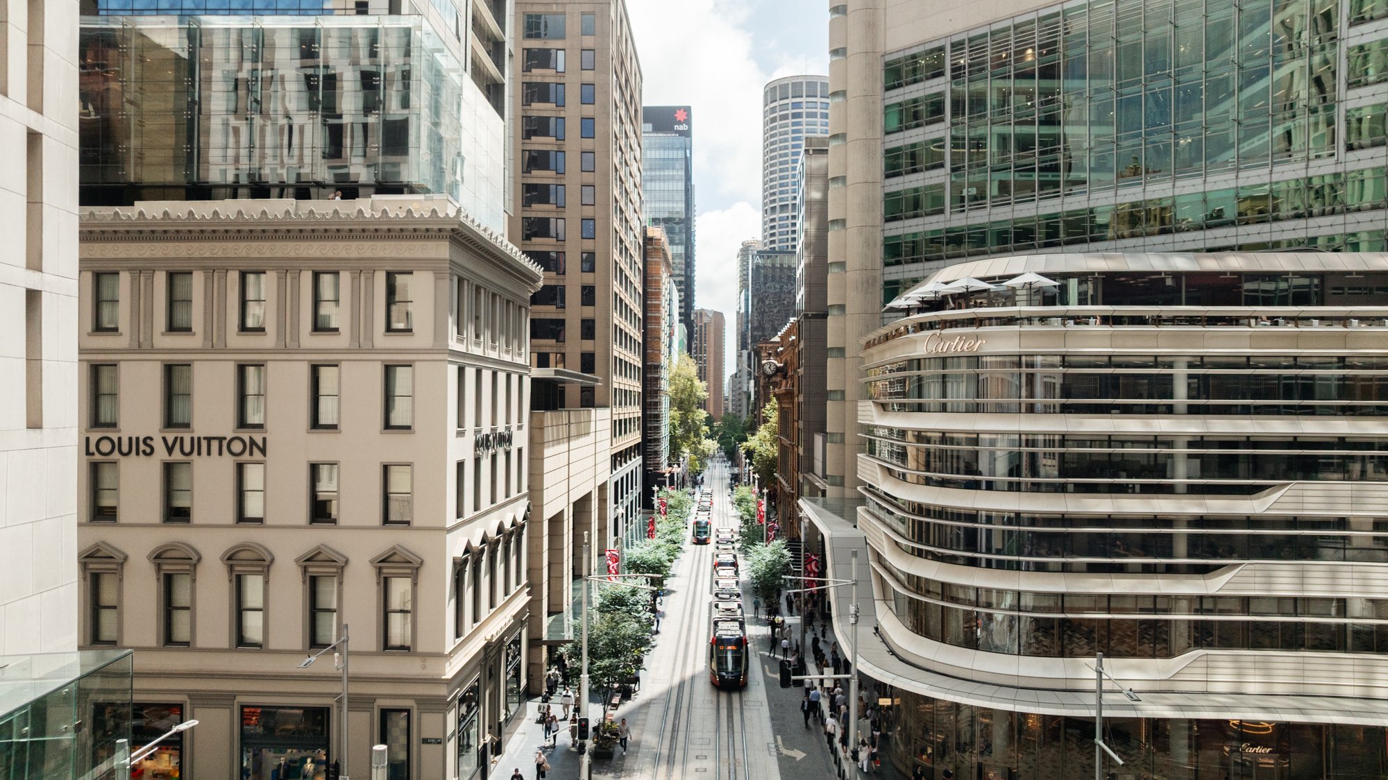 A busy city street with tall buildings, shops, and pedestrians, overhead view of a modern downtown area. Designed by fjcStudio Photo by Ruth Gold