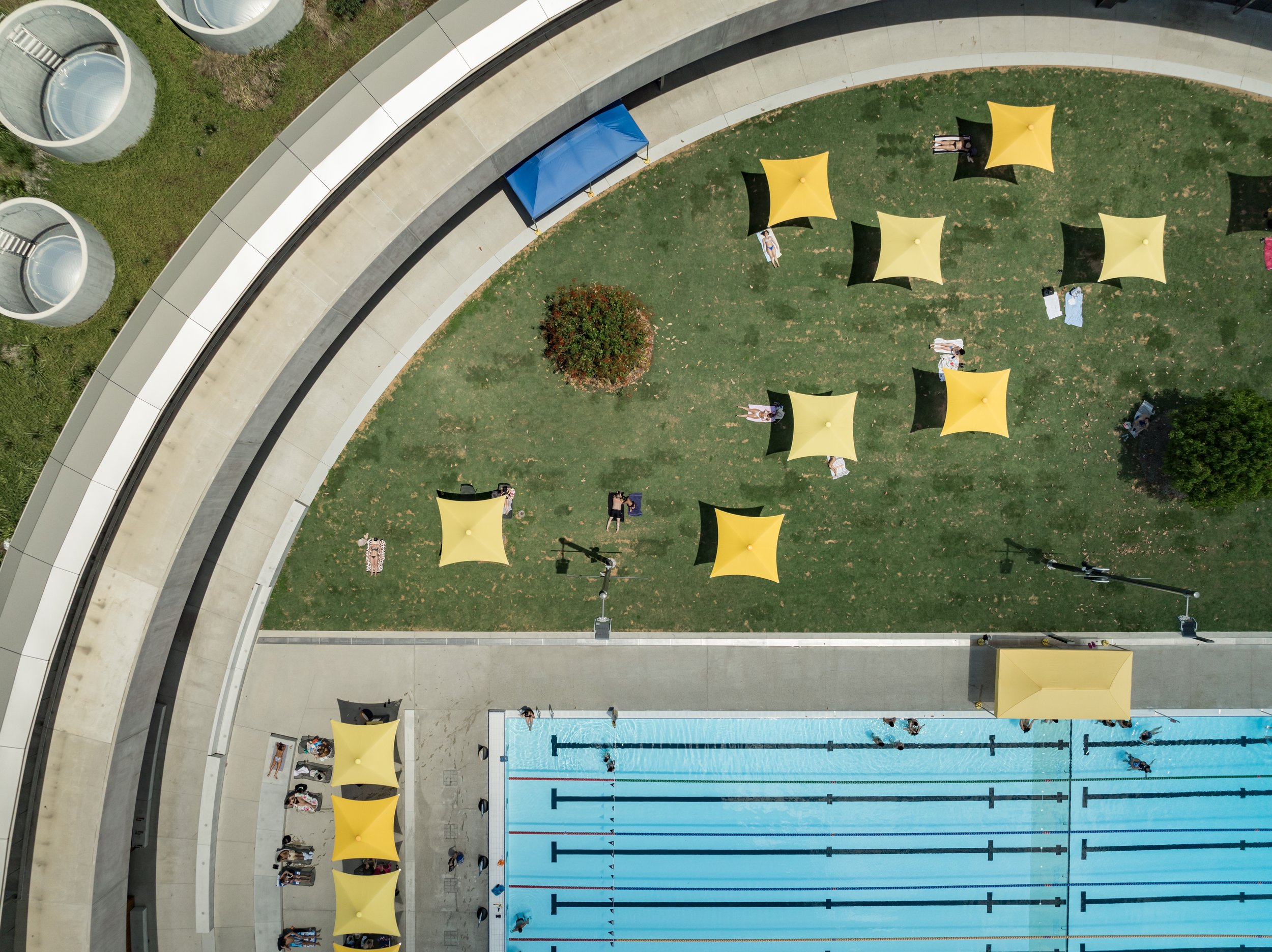 Aerial view of an outdoor pool area with swimmers, yellow tents, and people relaxing on the grass and around the pool. Designed by McGregor Coxall Photo by Ruth Gold