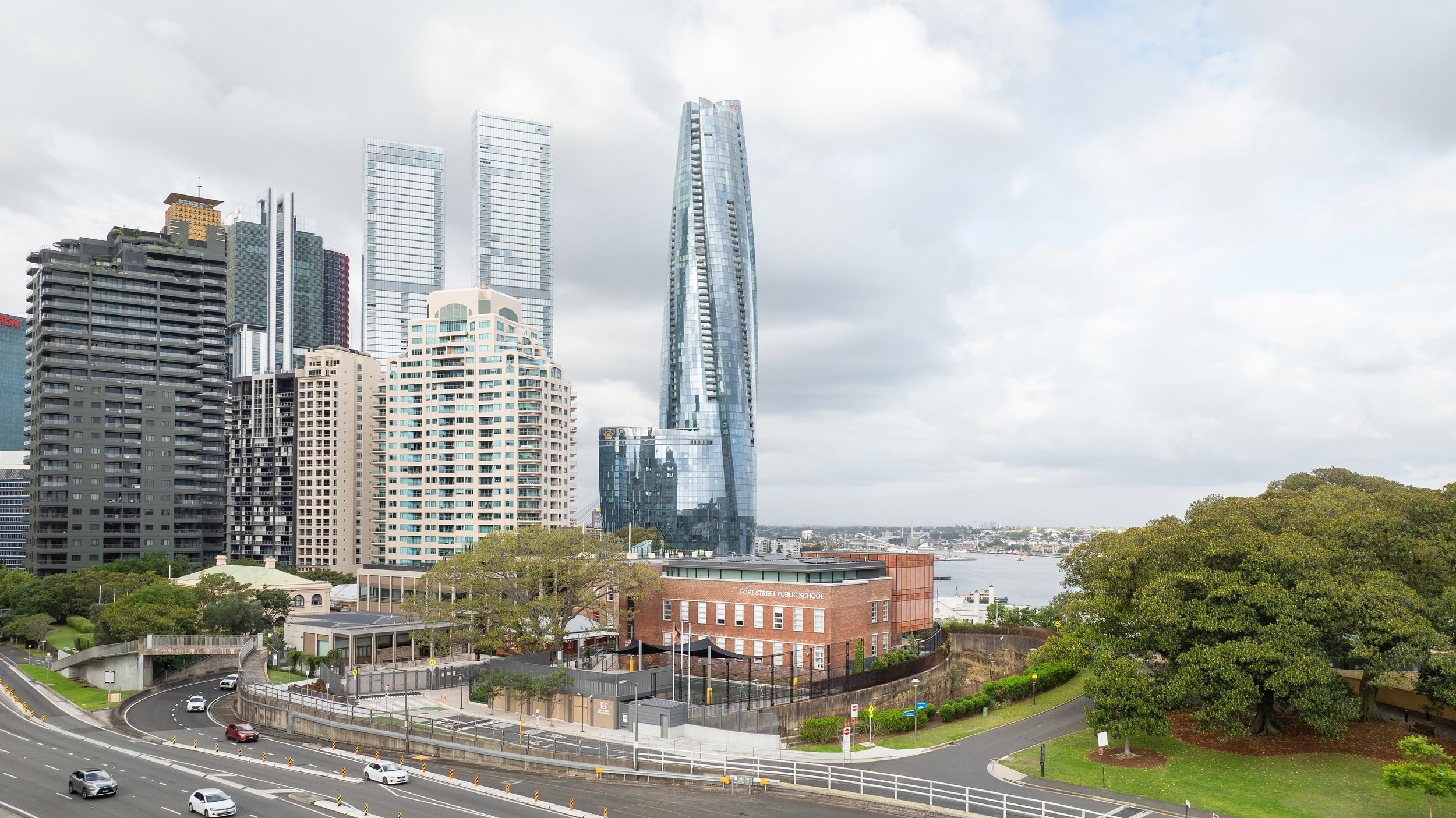 City skyline with tall modern skyscrapers, a school building labeled Ford Street Public School, a river in the background, trees, and a street with cars. Designed by fjcStudio Photo by Ruth Gold
