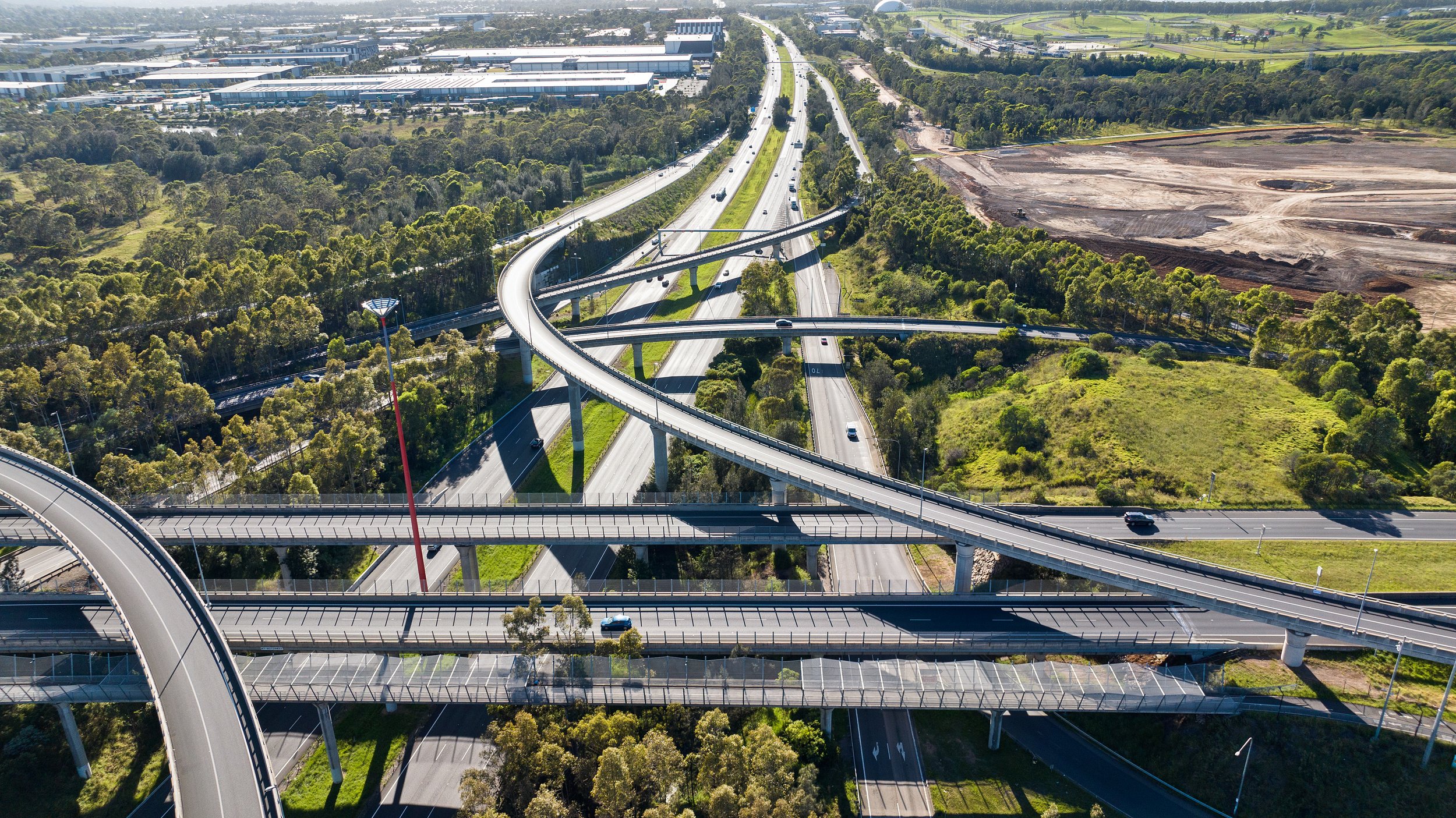 Aerial view of multiple interconnected highway overpasses and ramps surrounded by trees, greenery, and some industrial buildings in the distance. Designed by Colin Polwarth Studio Photo by Ruth Gold