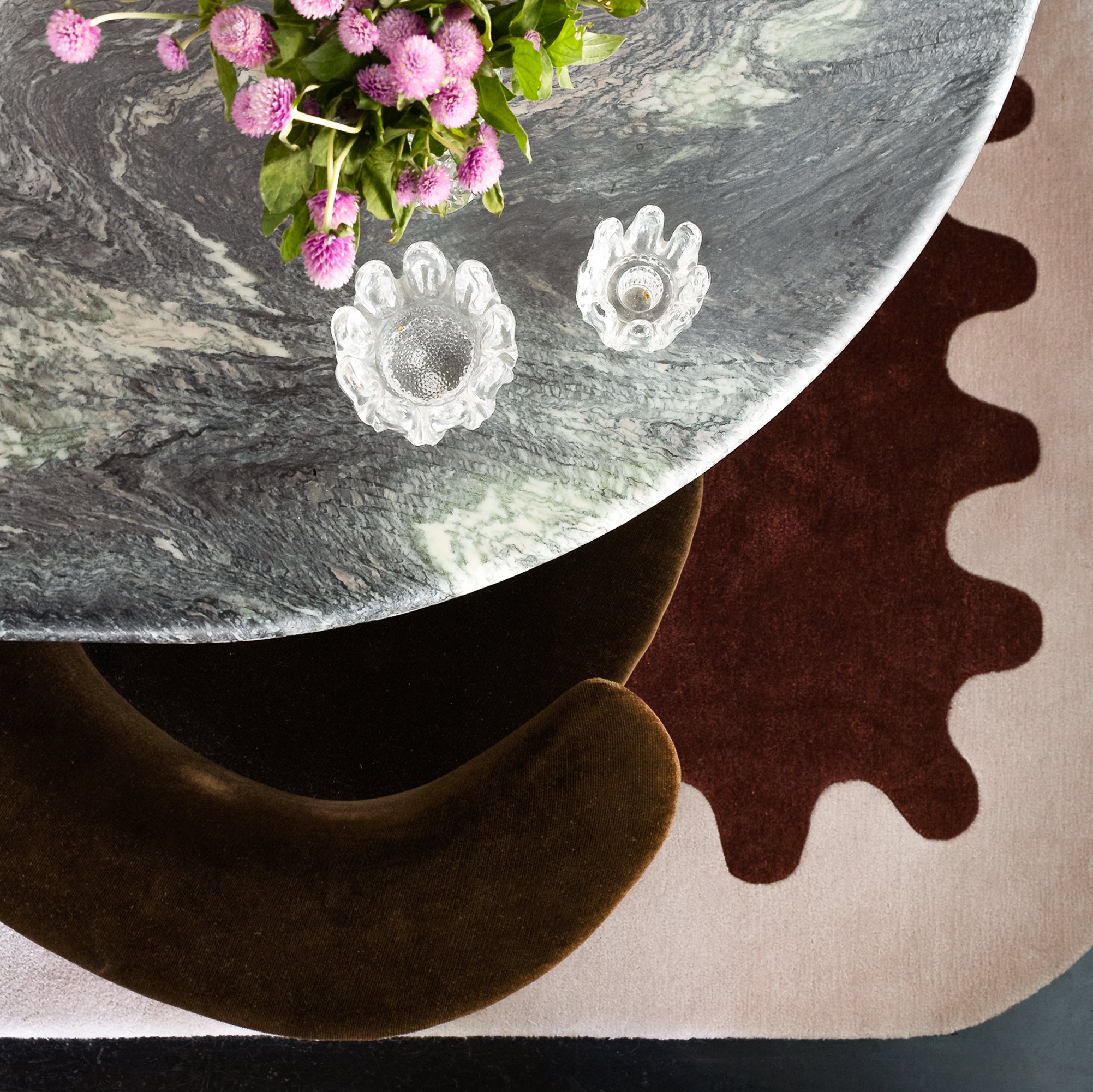 Top view of a round marble table with a pink flower arrangement and two small glass candle holders, one with a candle. The table is over a dark brown velvet chair, and beneath the chair is a decorative rug with a scalloped edge design. SMAC Studio Ph
