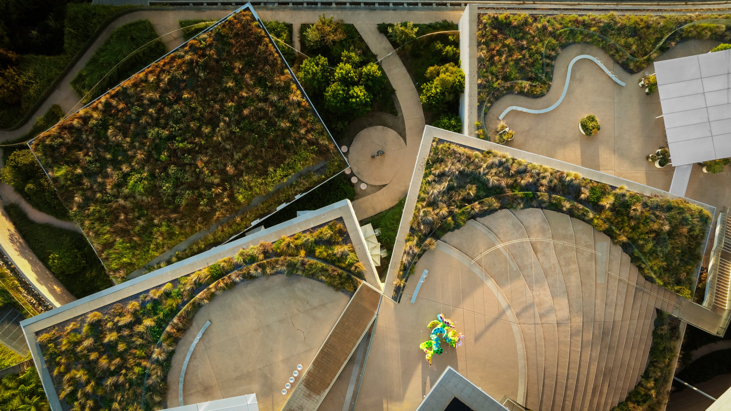 An aerial view of a modern building complex with rooftop gardens, pathways, and a paved open area with colorful sculptures. Designed by McGregor Coxall Photo by Ruth Gold