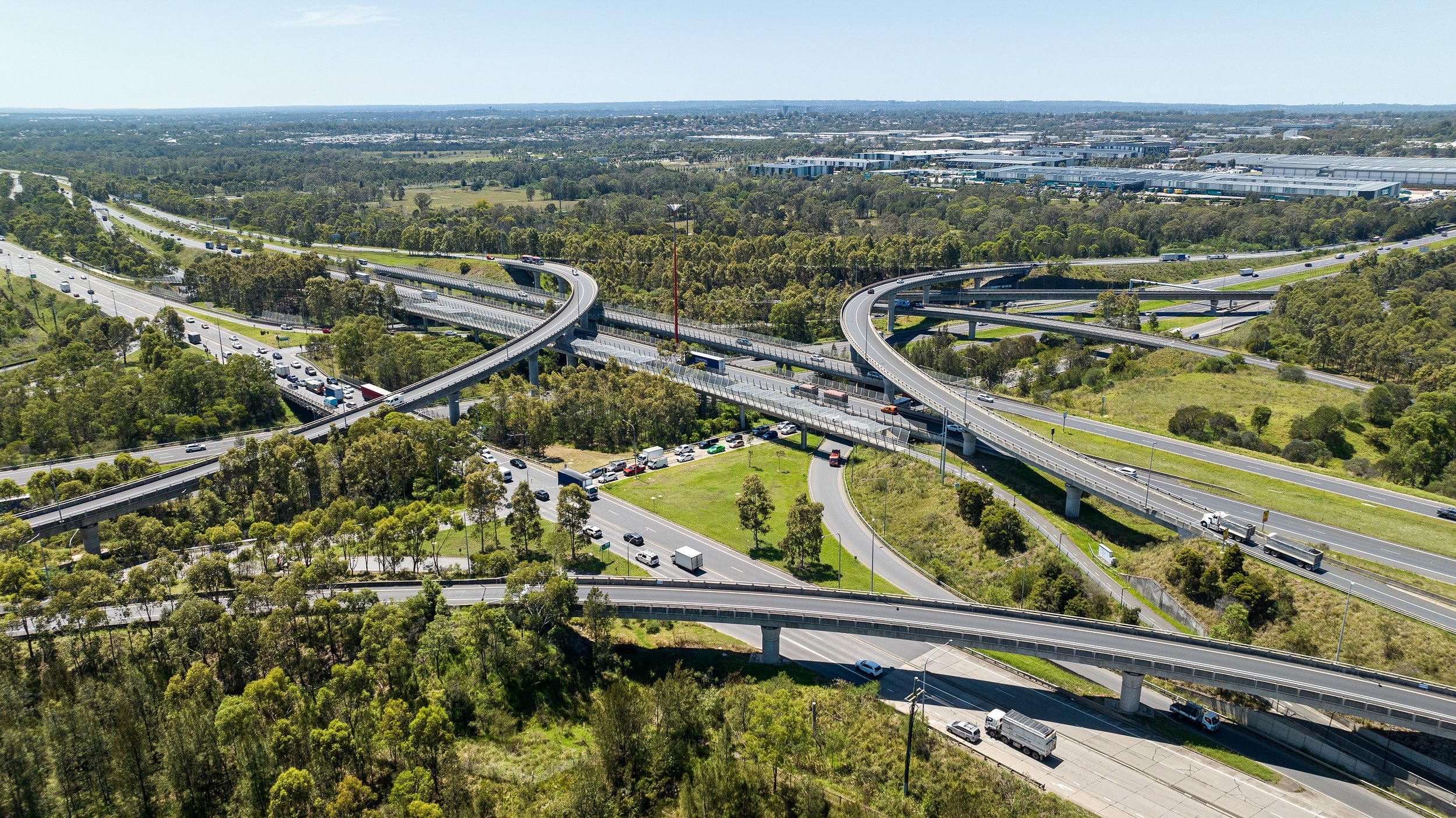 Aerial view of a complex highway interchange with multiple overpasses and ramps, surrounded by green trees and some commercial buildings in the distance. Designed by Colin Polwarth Studio Photo by Ruth Gold