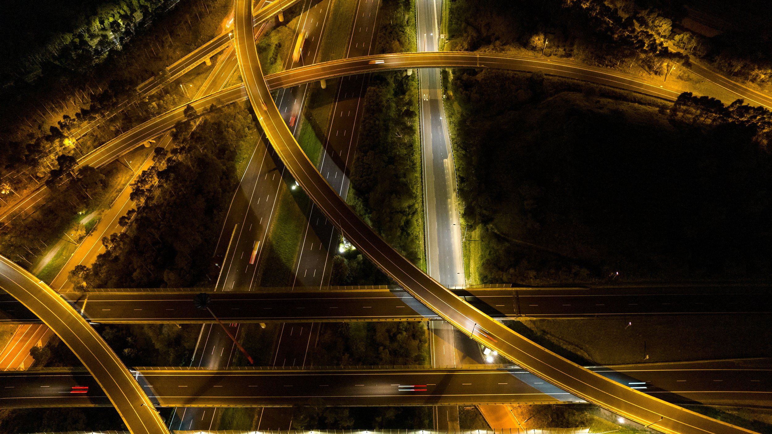 Nighttime aerial view of a multi-level highway interchange with several overpasses illuminated by streetlights, surrounded by trees and dark areas. Designed by Colin Polwarth Studio Photo by Ruth Gold