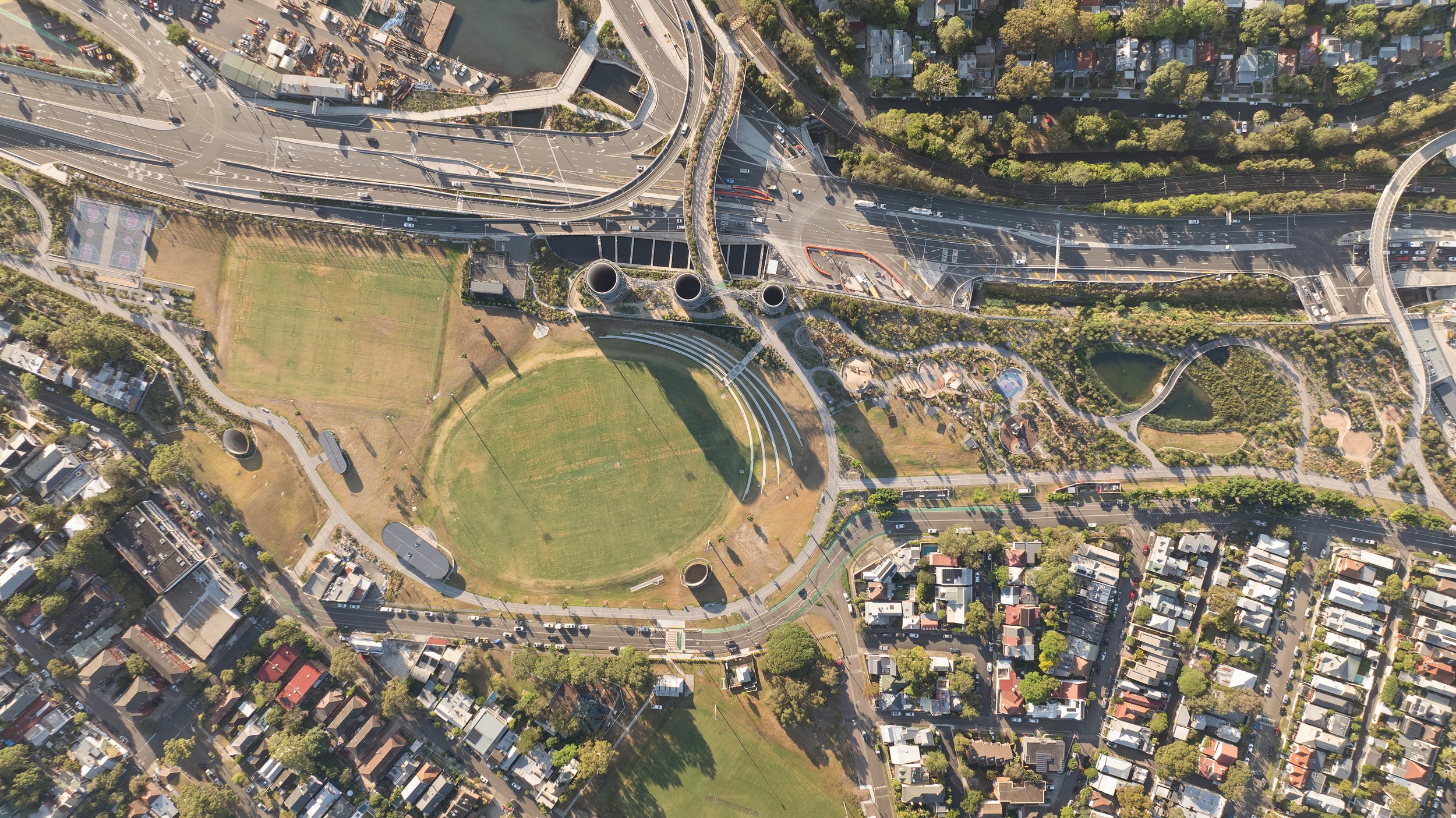 Aerial view of a park with sports fields, winding pathways, and small water features, surrounded by roads and residential neighborhoods. Designed by McGregor Coxall Photo by Ruth Gold