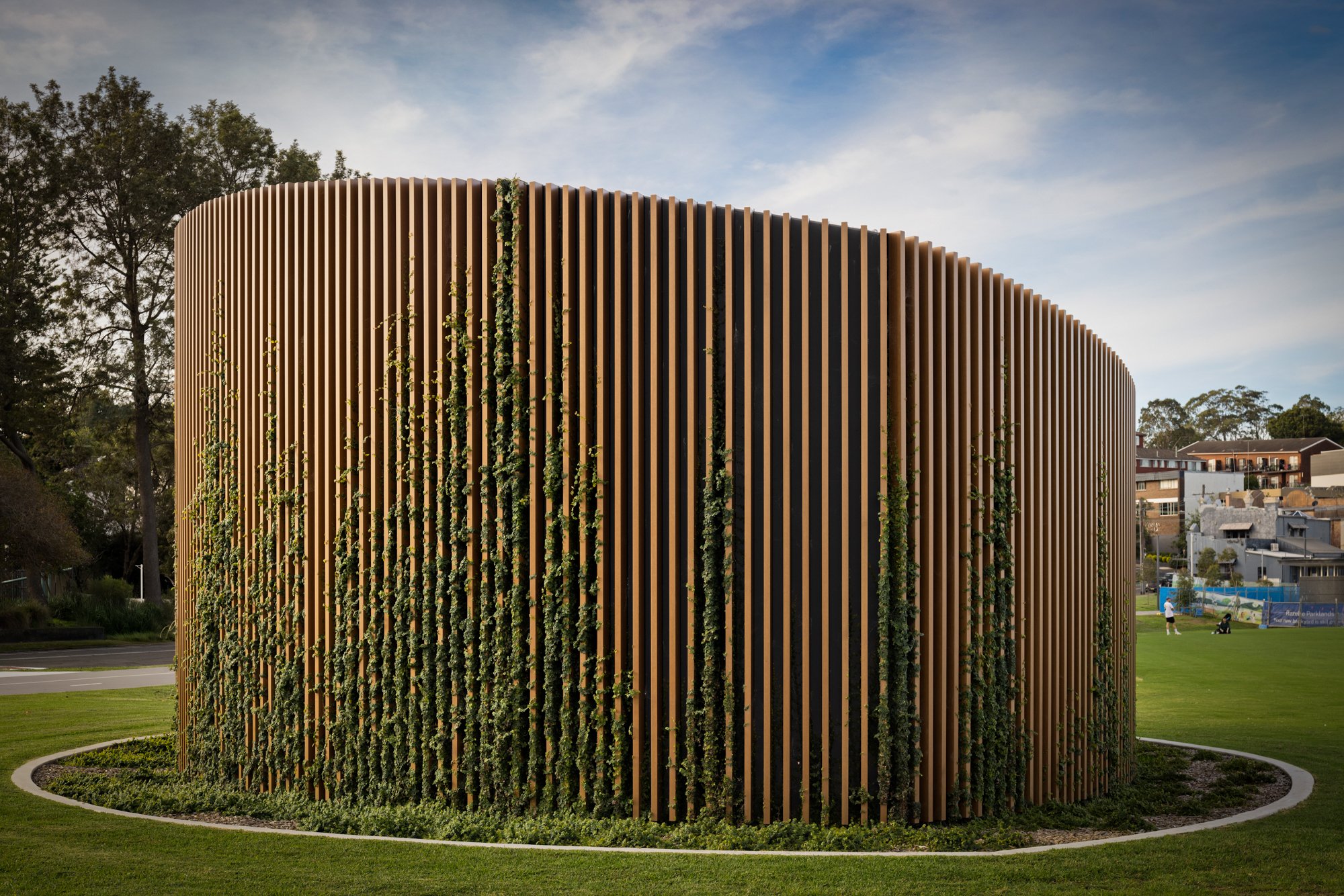 A modern, circular wooden building with vertical slats and green vines growing on the exterior, surrounded by a grassy area and trees, under a partly cloudy sky. Designed by Colin Polwarth Studio Photo by Ruth Gold