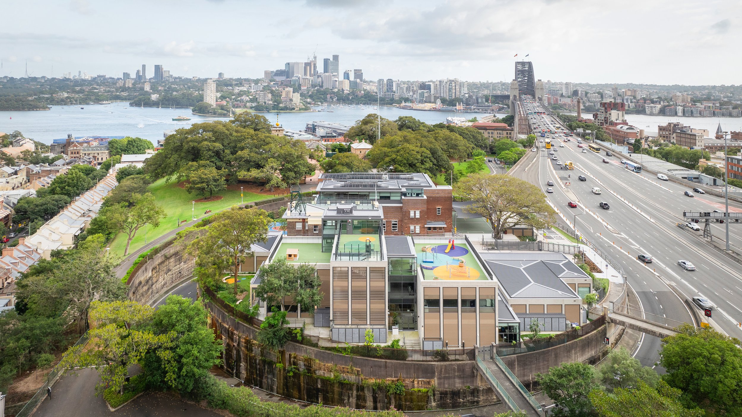 A cityscape with a river, high-rise buildings, and a bridge. In the foreground, an urban school or community center with sports courts and green space, surrounded by trees and adjacent to a highway. Designed by fjcStudio Photo by Ruth Gold