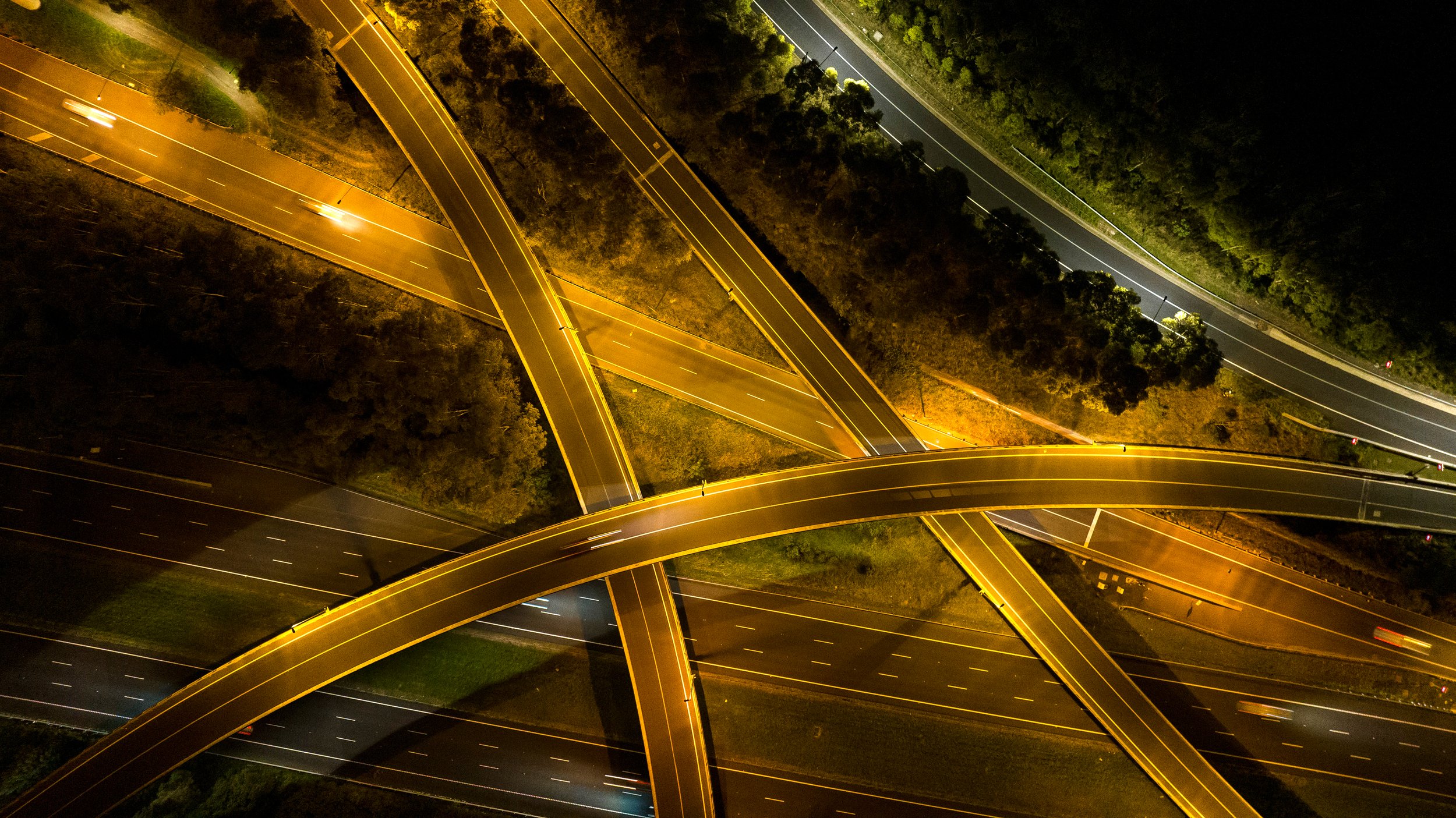 Night aerial view of a complex highway interchange with multiple overpasses illuminated by streetlights. Designed by Colin Polwarth Studio Photo by Ruth Gold