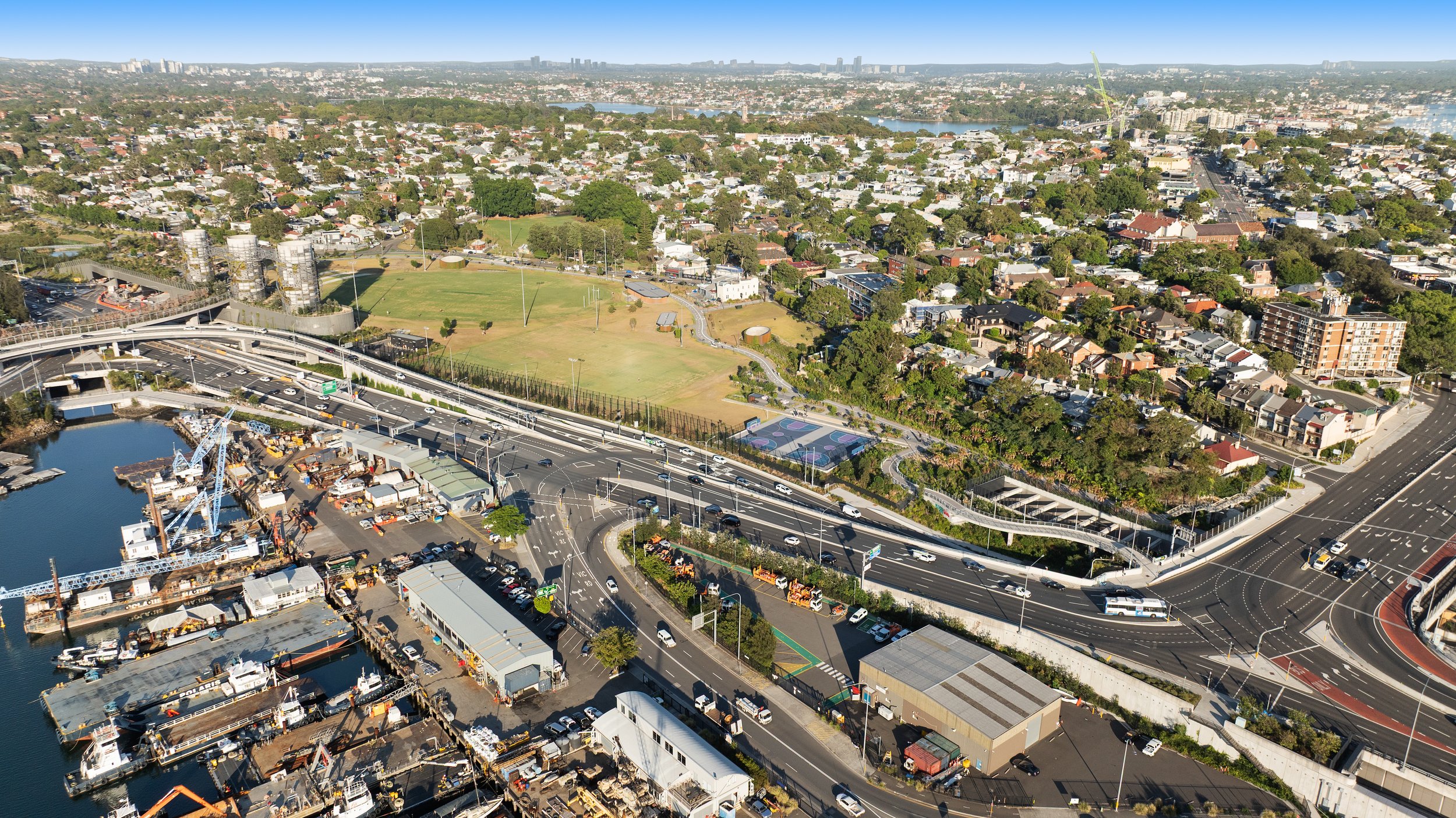 Aerial view of a cityscape featuring a marina with boats, a construction site with cranes, a highway with multiple lanes and cars, a park with a playground, residential neighborhoods, and a distant city skyline with high-rise buildings. Designed by M