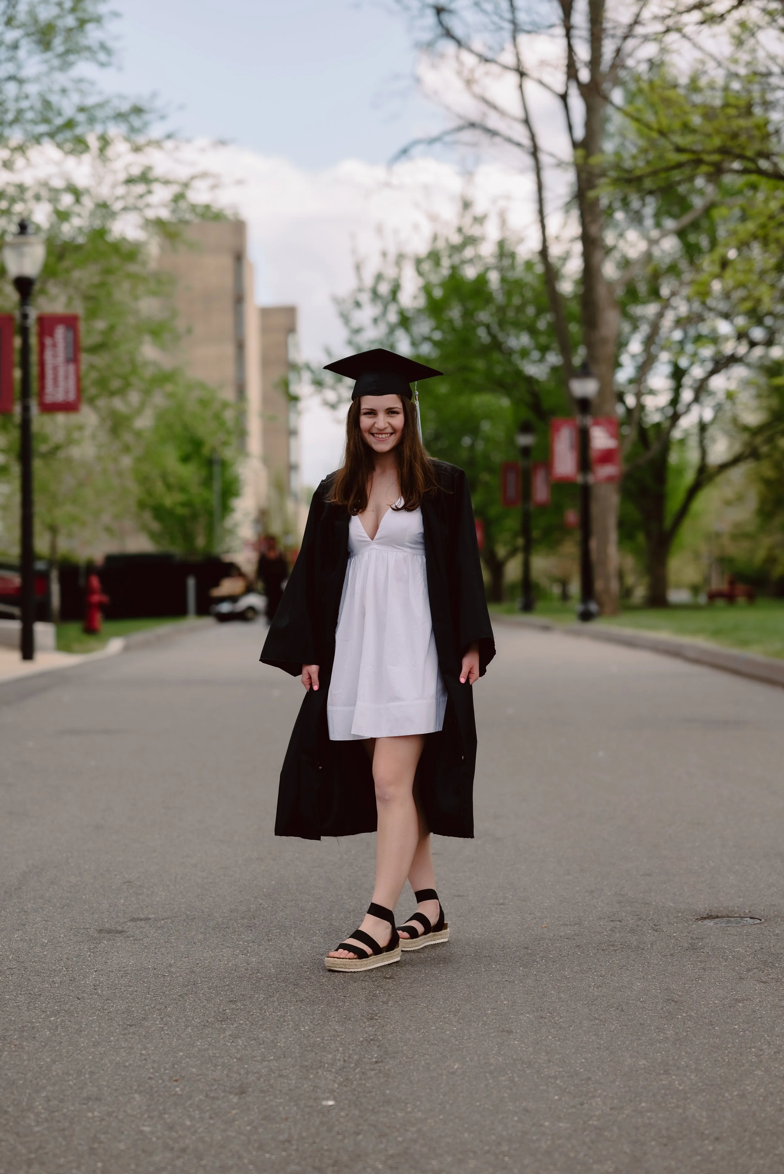 A young woman in graduation attire, including a black cap and gown with a white dress underneath, smiling and standing on an empty street with trees and buildings in the background.
