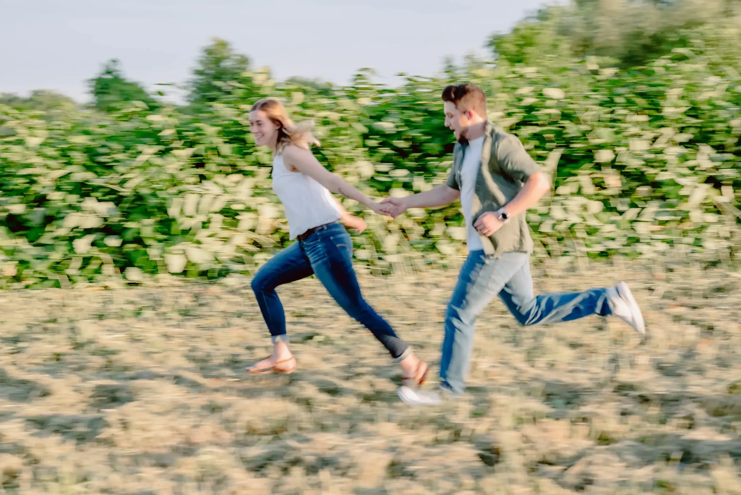 A young couple running and holding hands through a field with green plants in the background.