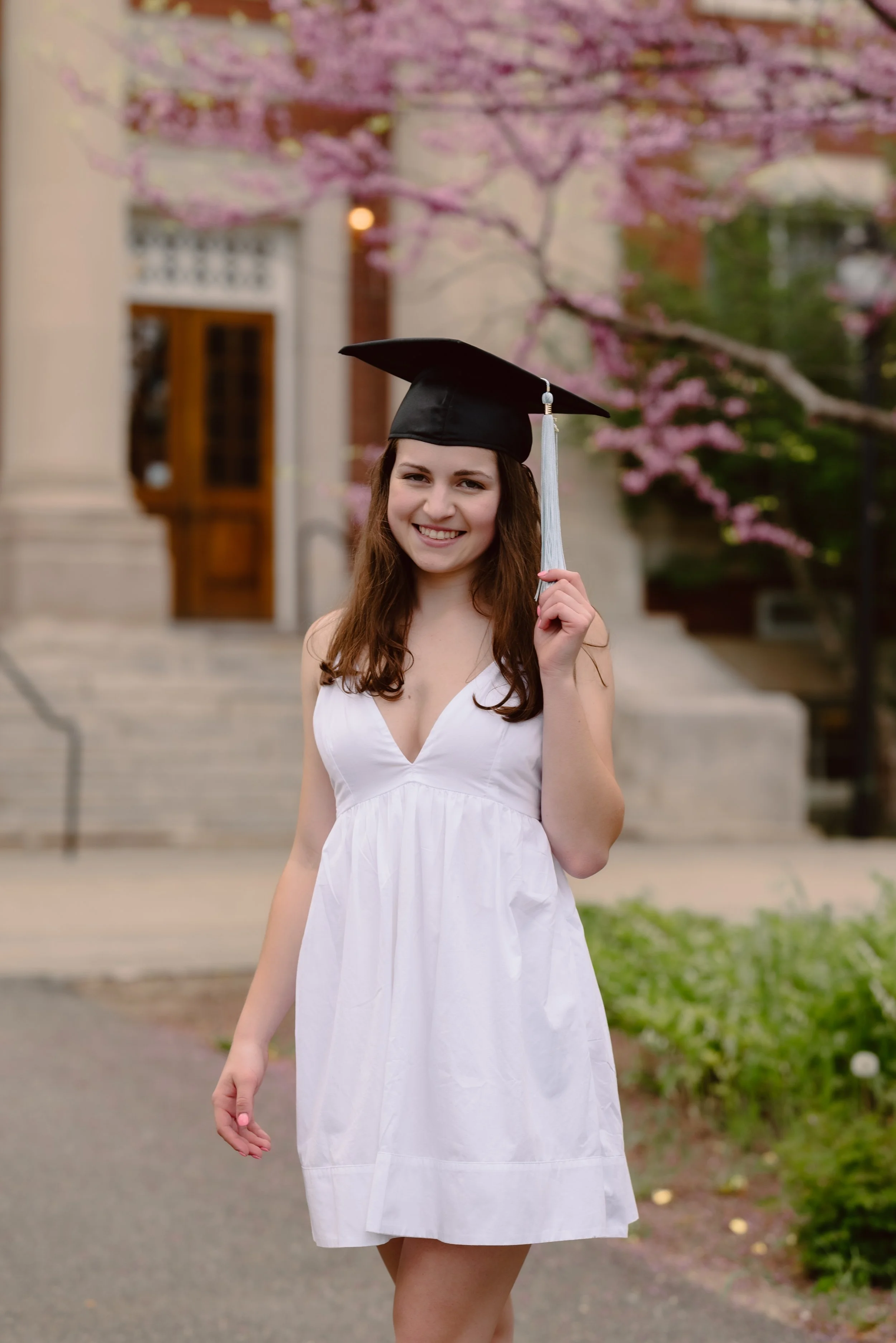 A young woman in a white dress holding a graduation cap outdoors with pink blossoming trees and a building in the background.