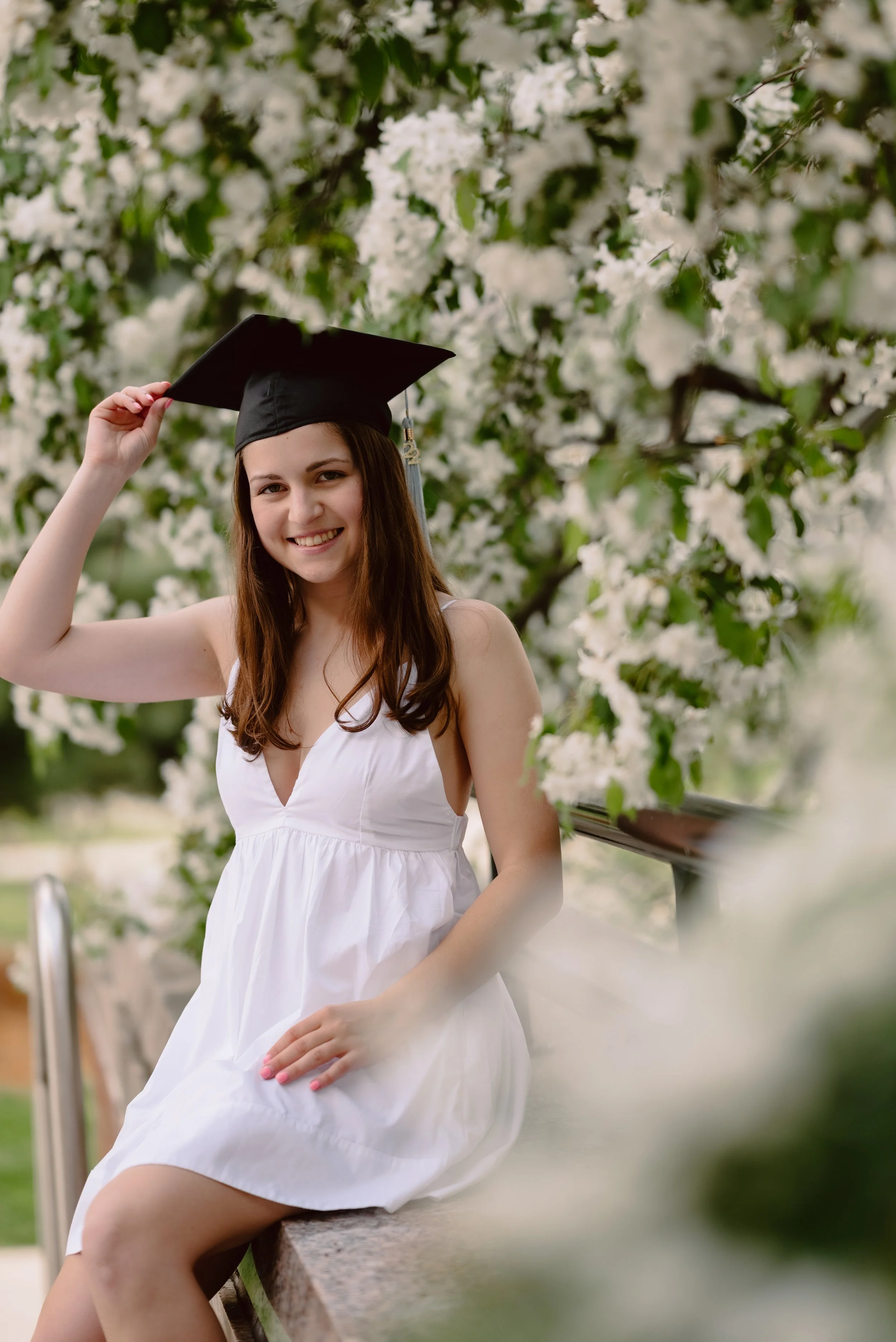 A young woman in a white dress and graduation cap sitting on a bench outdoors, smiling and adjusting her cap, surrounded by white flowering trees.