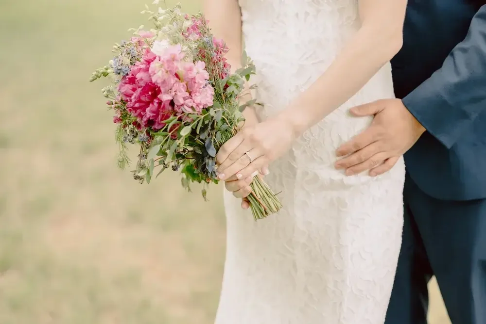 Close-up of a bride holding a bouquet of pink and purple flowers, with a groom standing next to her with his hand on her waist, during a wedding.