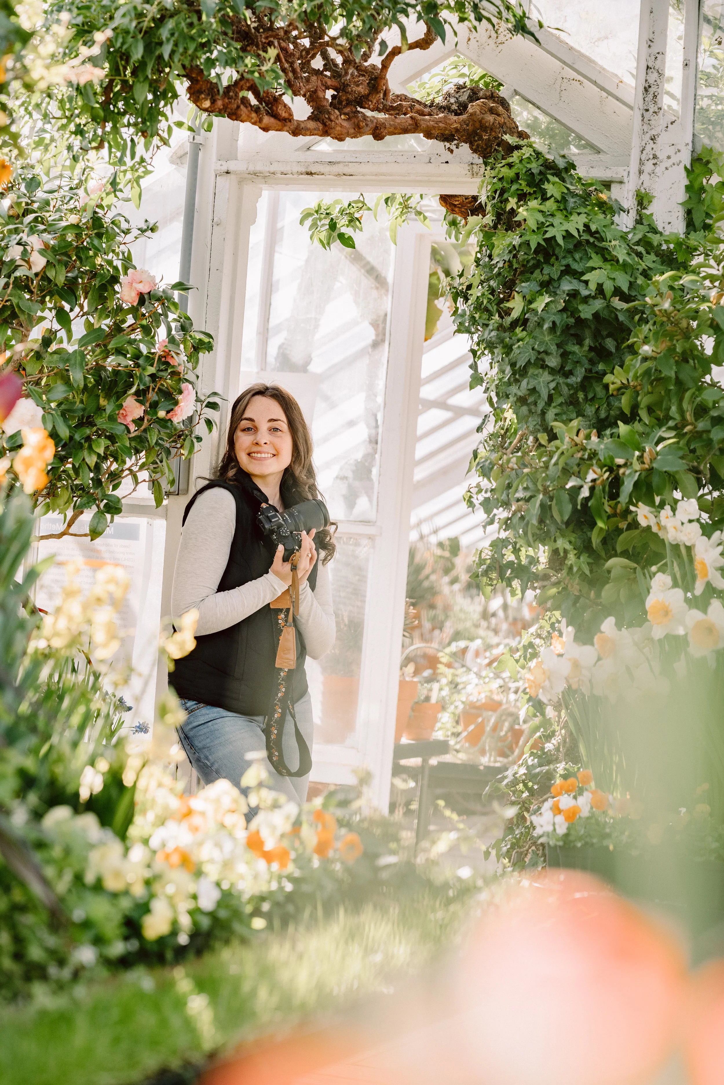 A woman smiling and holding a camera inside a greenhouse filled with various flowers and green plants.