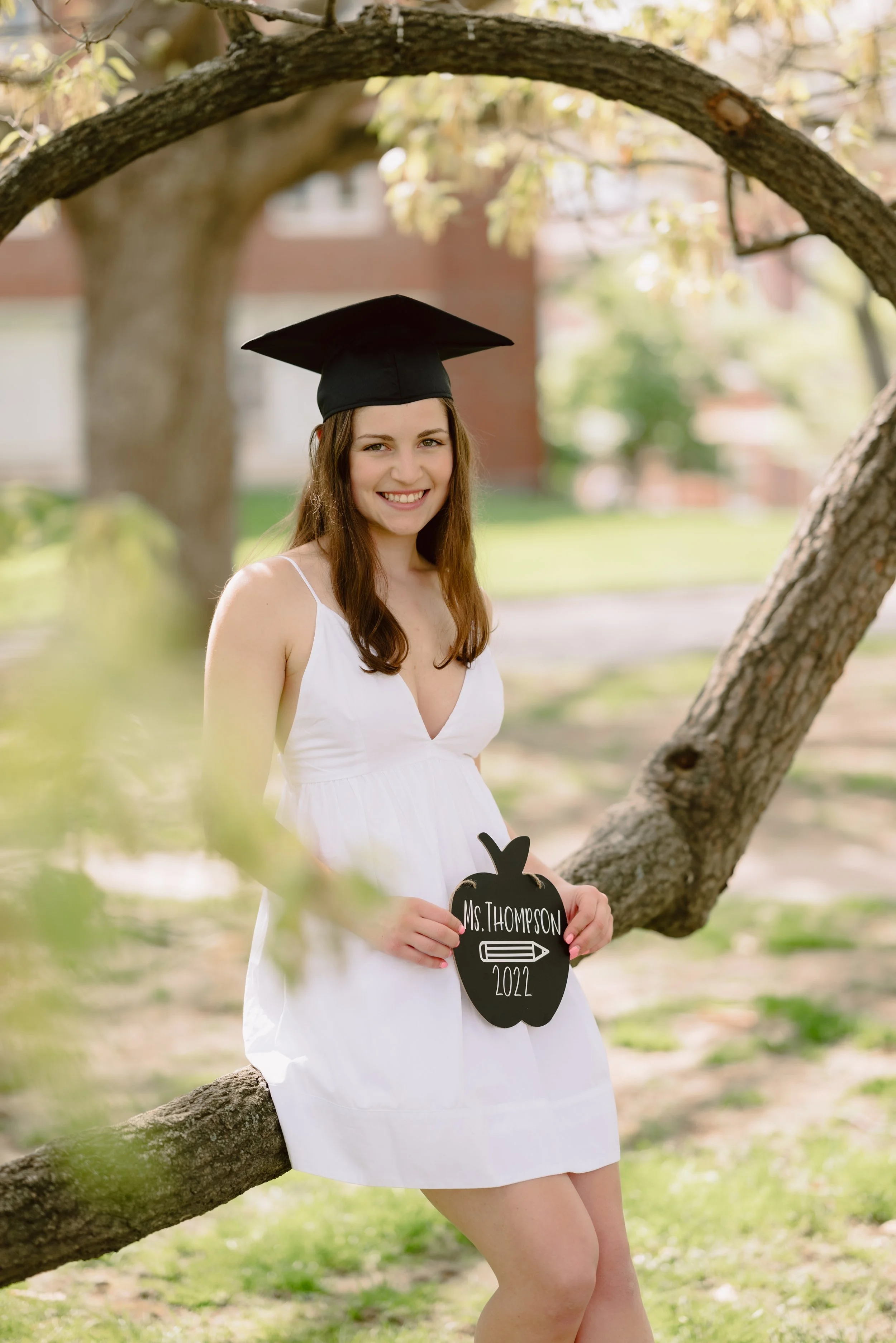 Young woman in a white dress and graduation cap smiling outdoors, holding a chalkboard sign shaped like a pumpkin with her name and graduation year written on it.