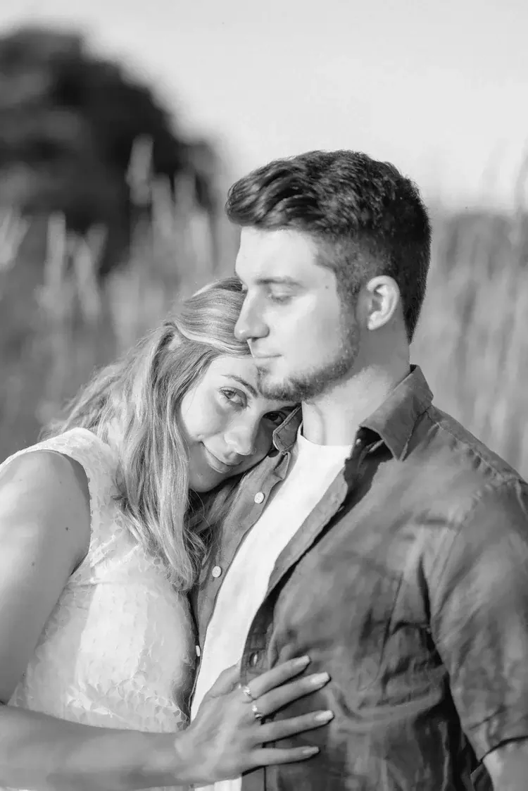 A black and white photo of a couple embracing outdoors, with the woman resting her head on the man's chest and both looking at the camera.