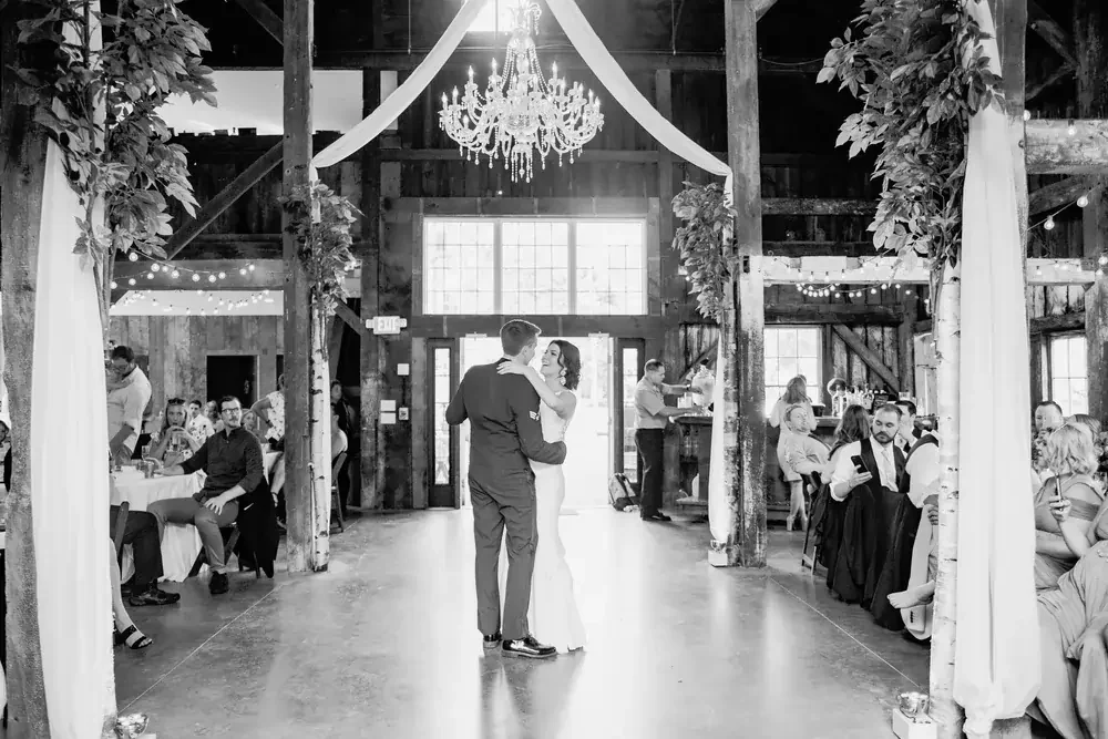 A couple dances in the center of a rustic wedding reception hall with wooden beams, string lights, and a chandelier.