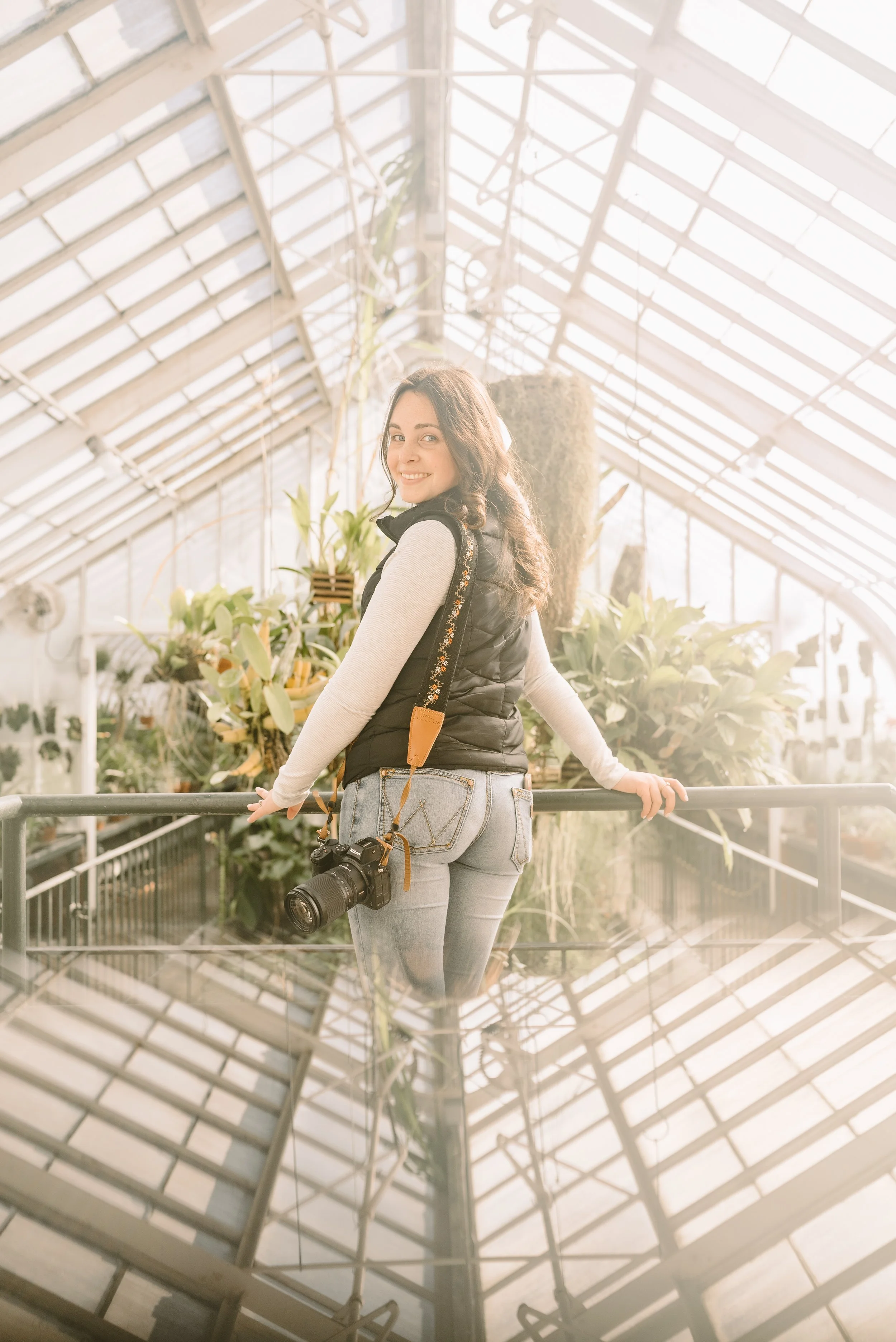 A woman with dark hair, wearing a black vest and light gray jeans, stands inside a greenhouse filled with plants. She is smiling at the camera, holding onto a black railing, and has a camera hanging from her neck. Sunlight streams through the glass c