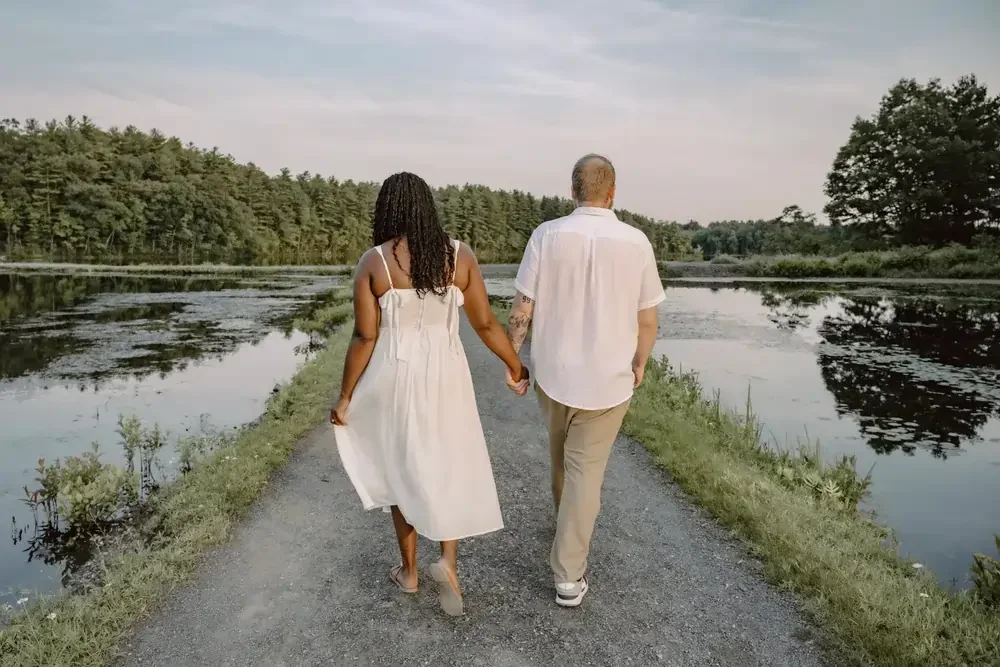 A couple walking hand in hand along a gravel path beside a calm body of water with trees in the background.