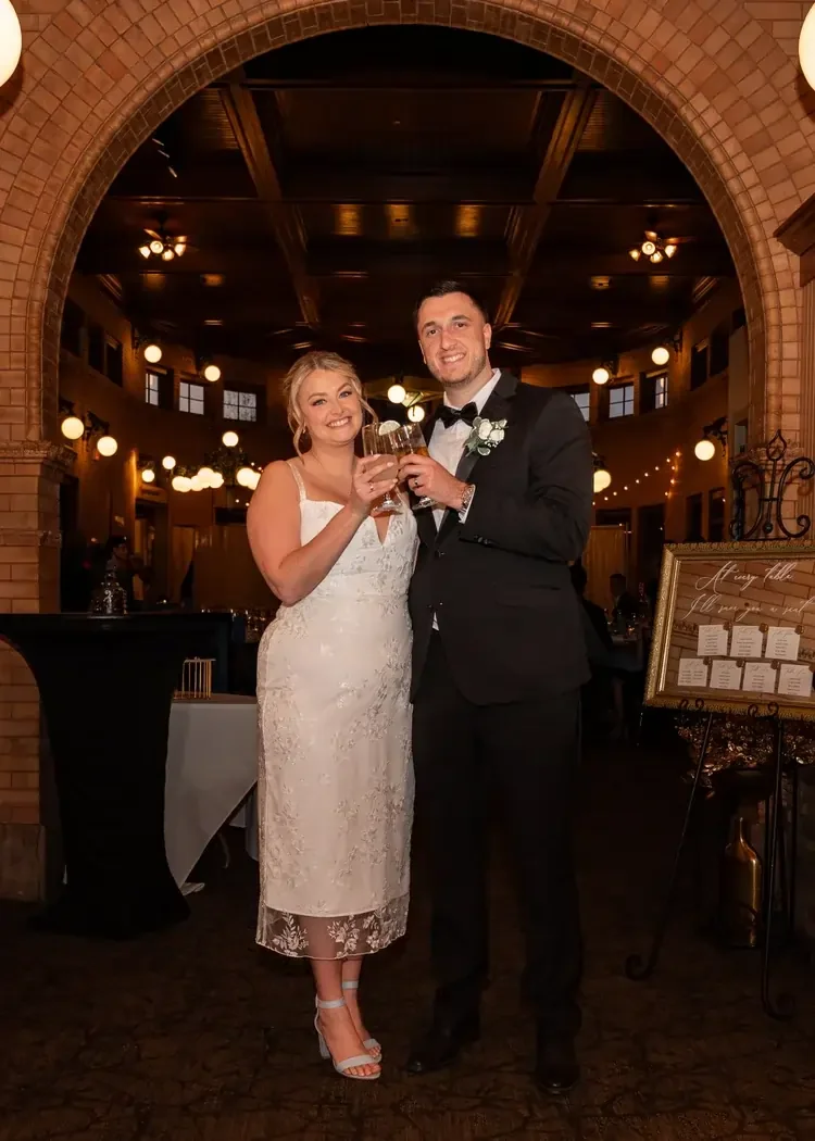 Bride and groom at their wedding reception, standing under an arched brick entrance, smiling and holding glasses of champagne.