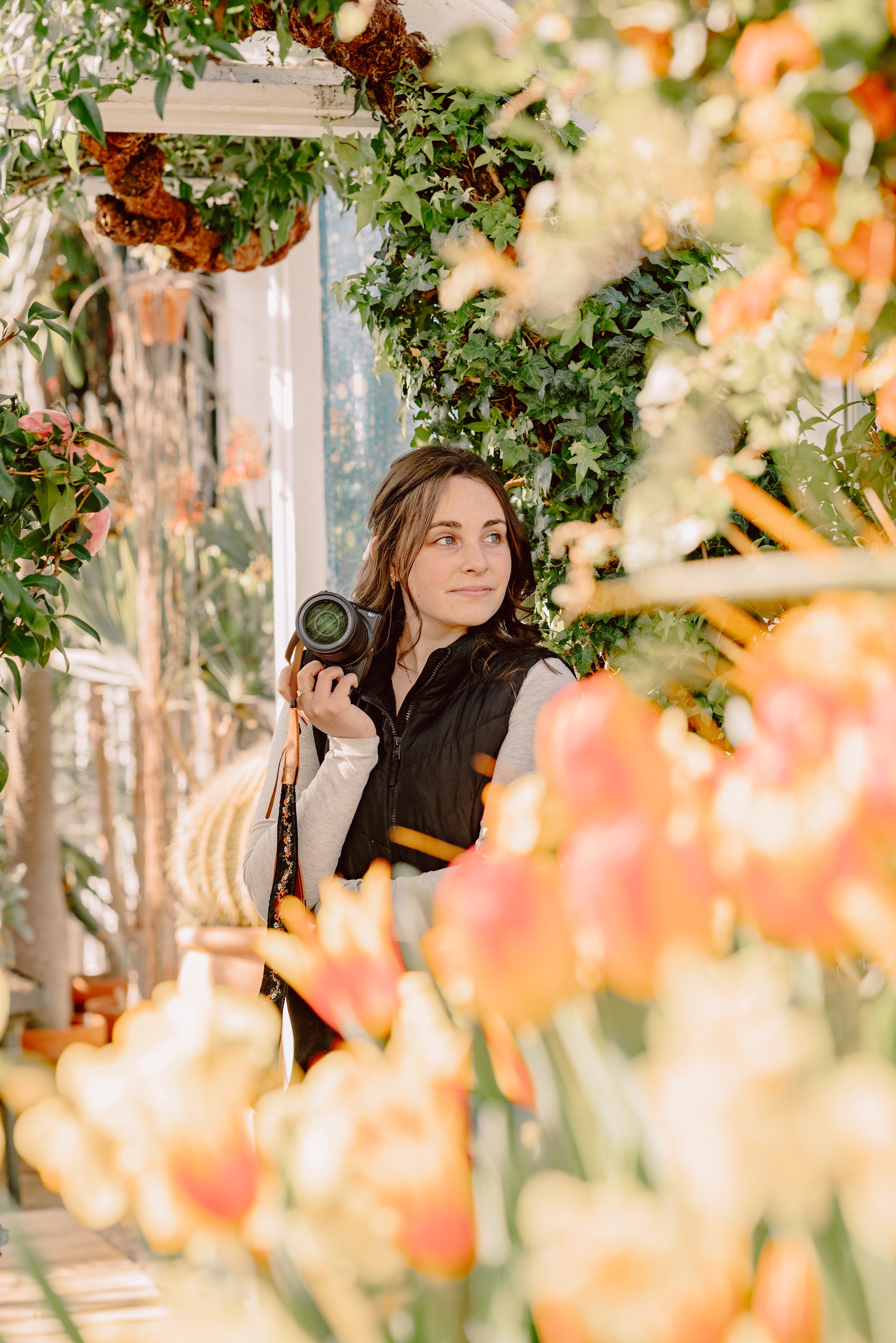 A woman holding a camera while standing among vibrant orange and yellow tulips in a garden or greenhouse with greenery.