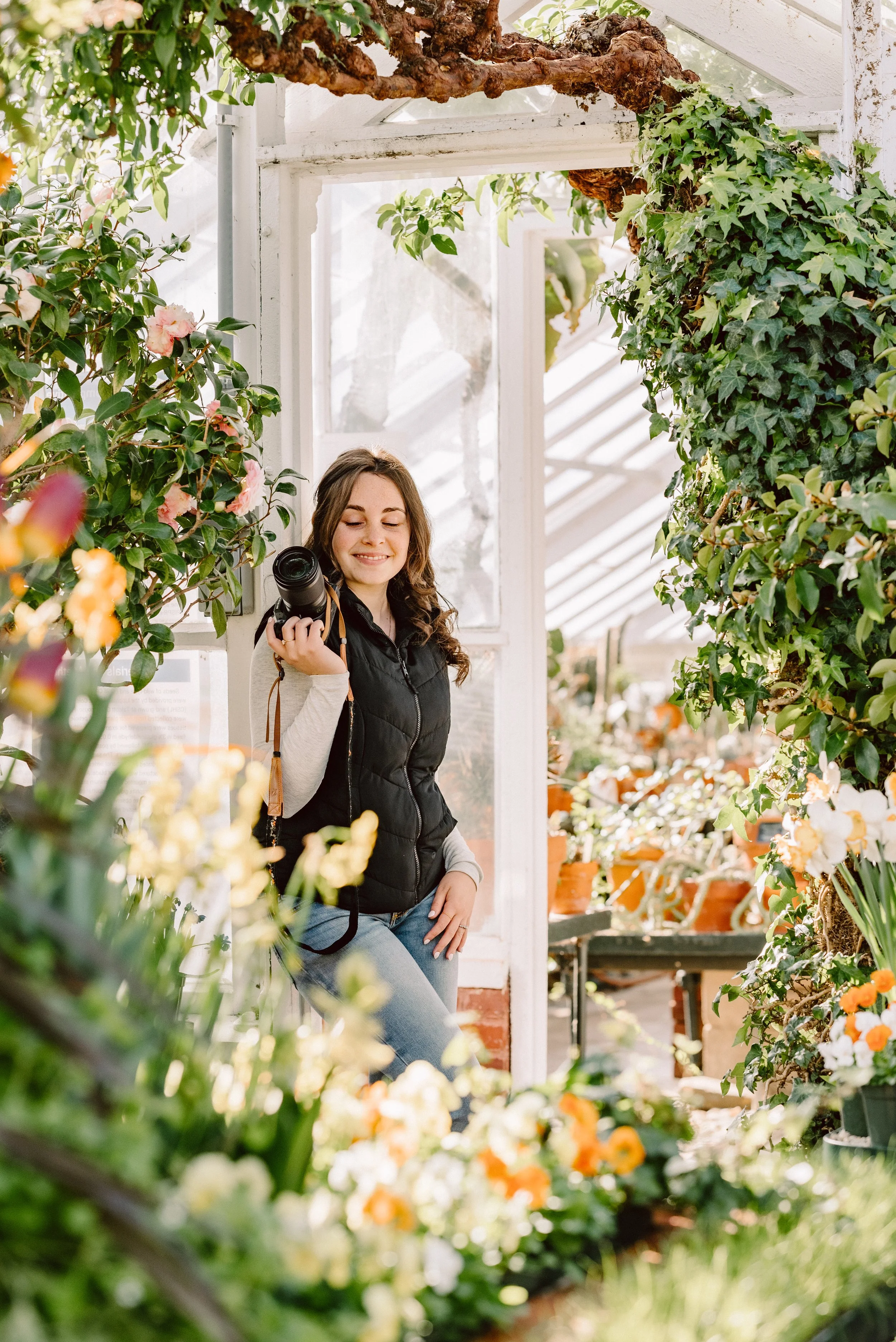 A young woman holding a camera inside a greenhouse, surrounded by blooming flowers and green plants.