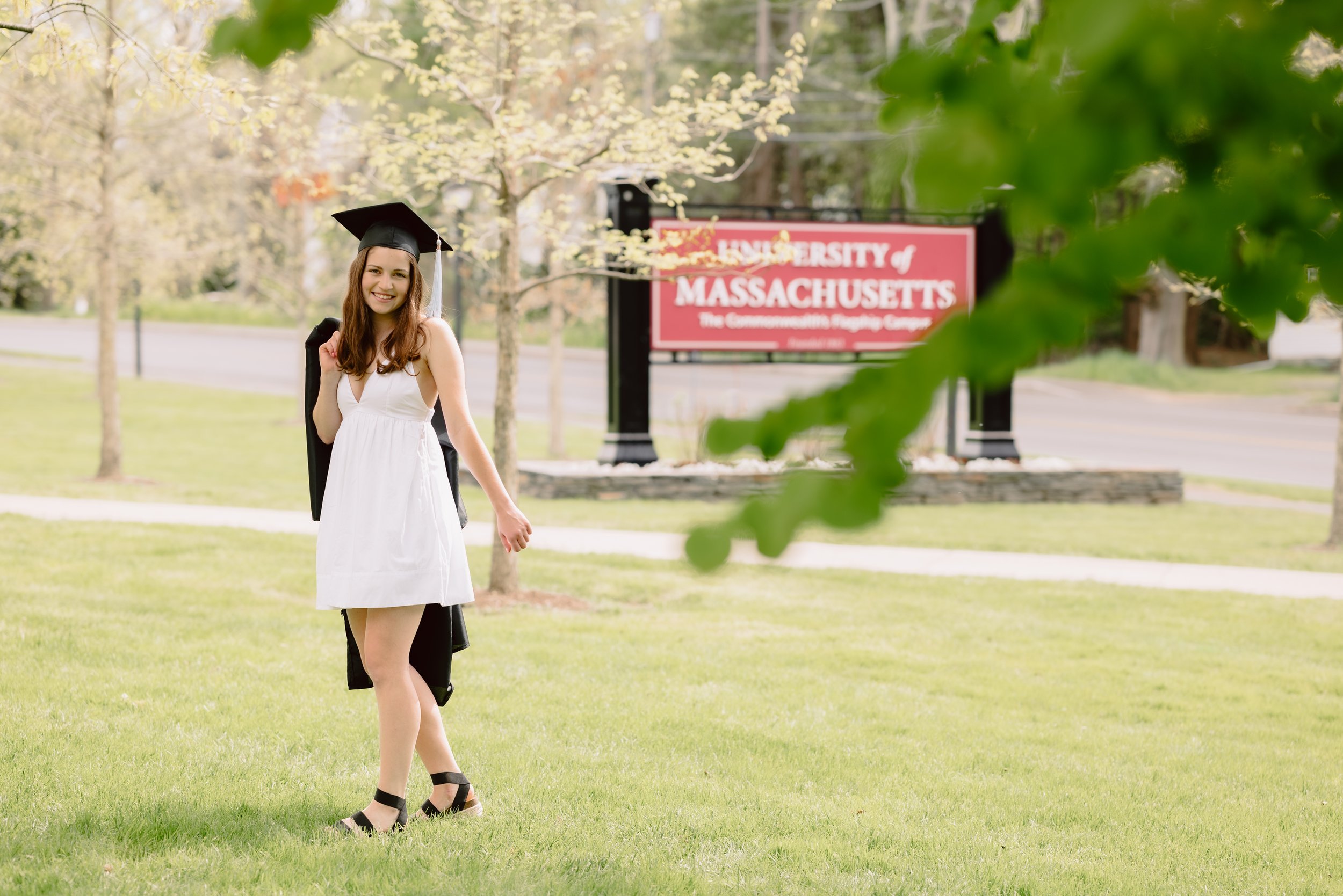 Young woman in a white dress and graduation cap standing on a grassy area, holding her gown with a university sign visible in the background.