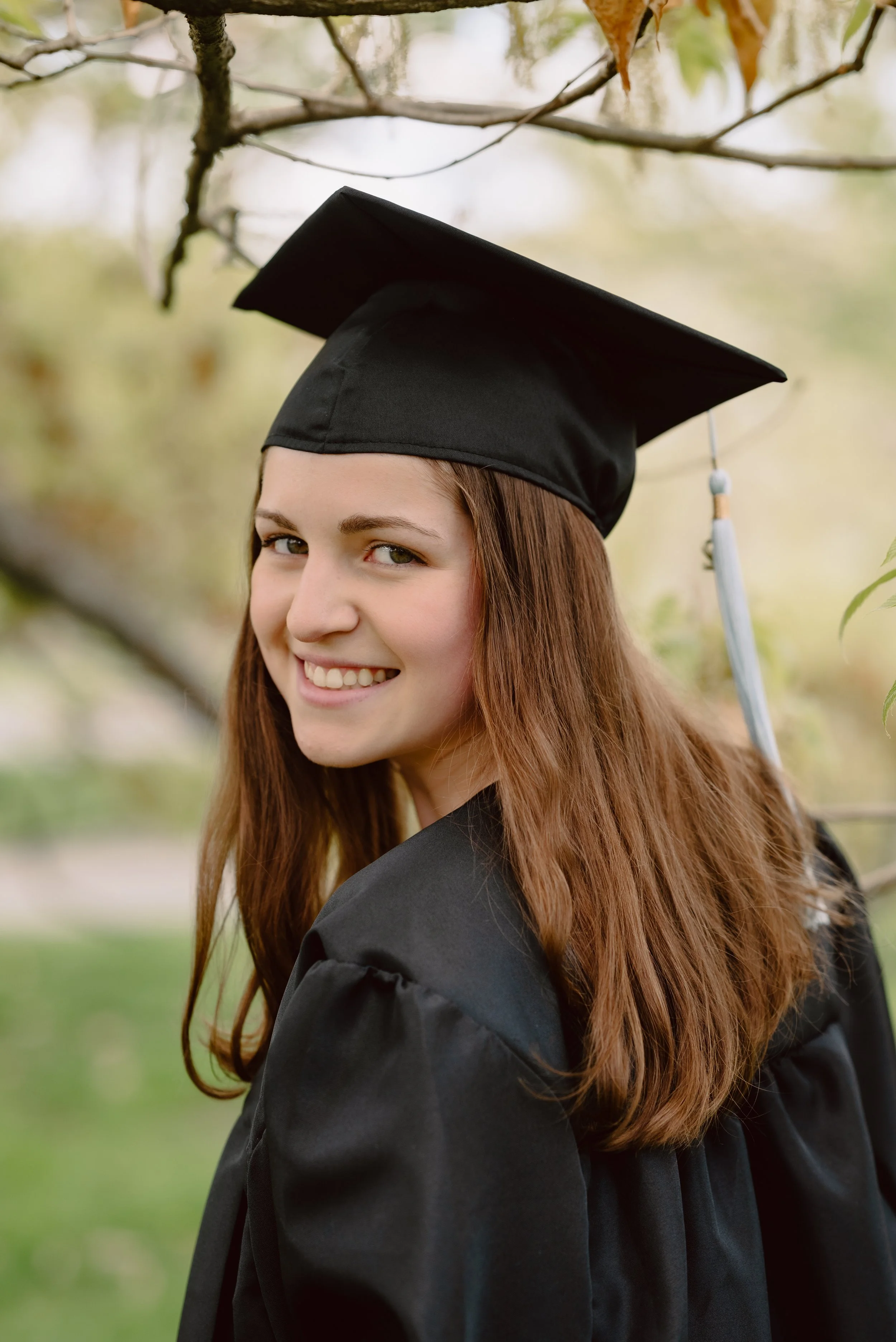 Young woman in a black graduation cap and gown smiling outdoors during daytime.
