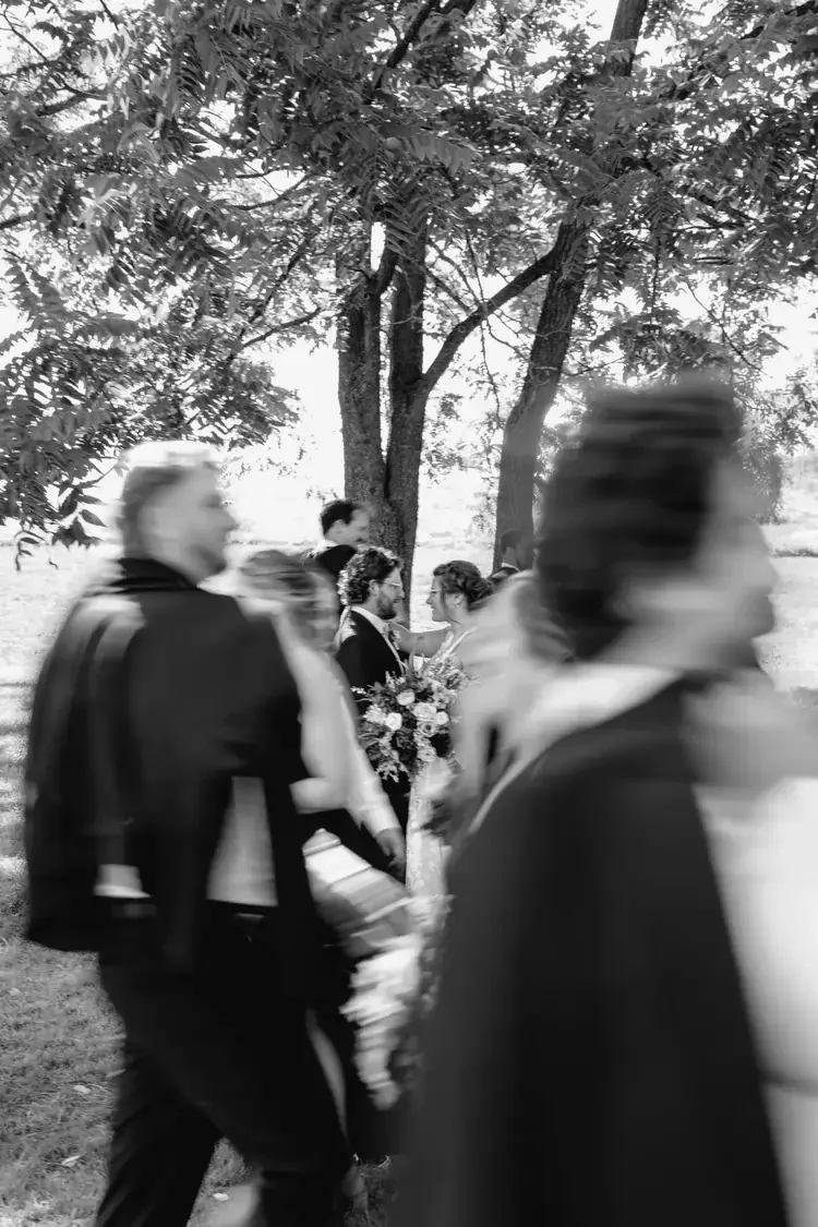 Blurred people walking past an outdoor wedding ceremony with a bride and groom standing under a tree, holding a bouquet of flowers.