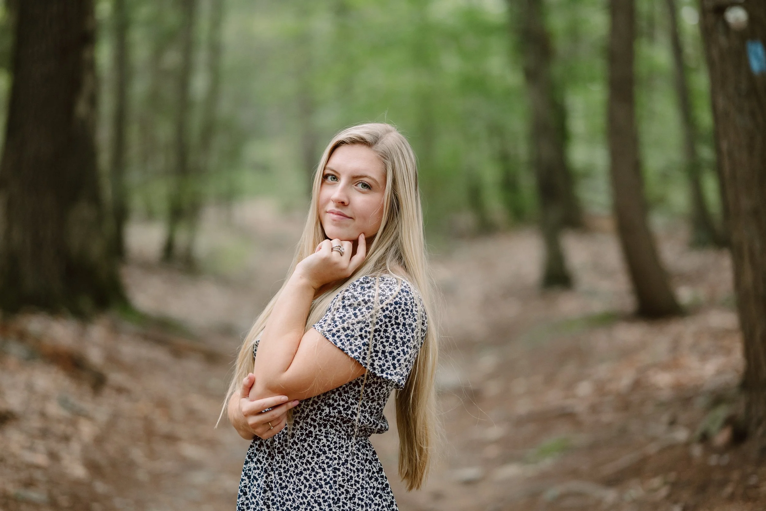 A young woman with long blonde hair in a patterned dress, standing in a wooded forest with green trees in the background, looking at the camera with a gentle smile.