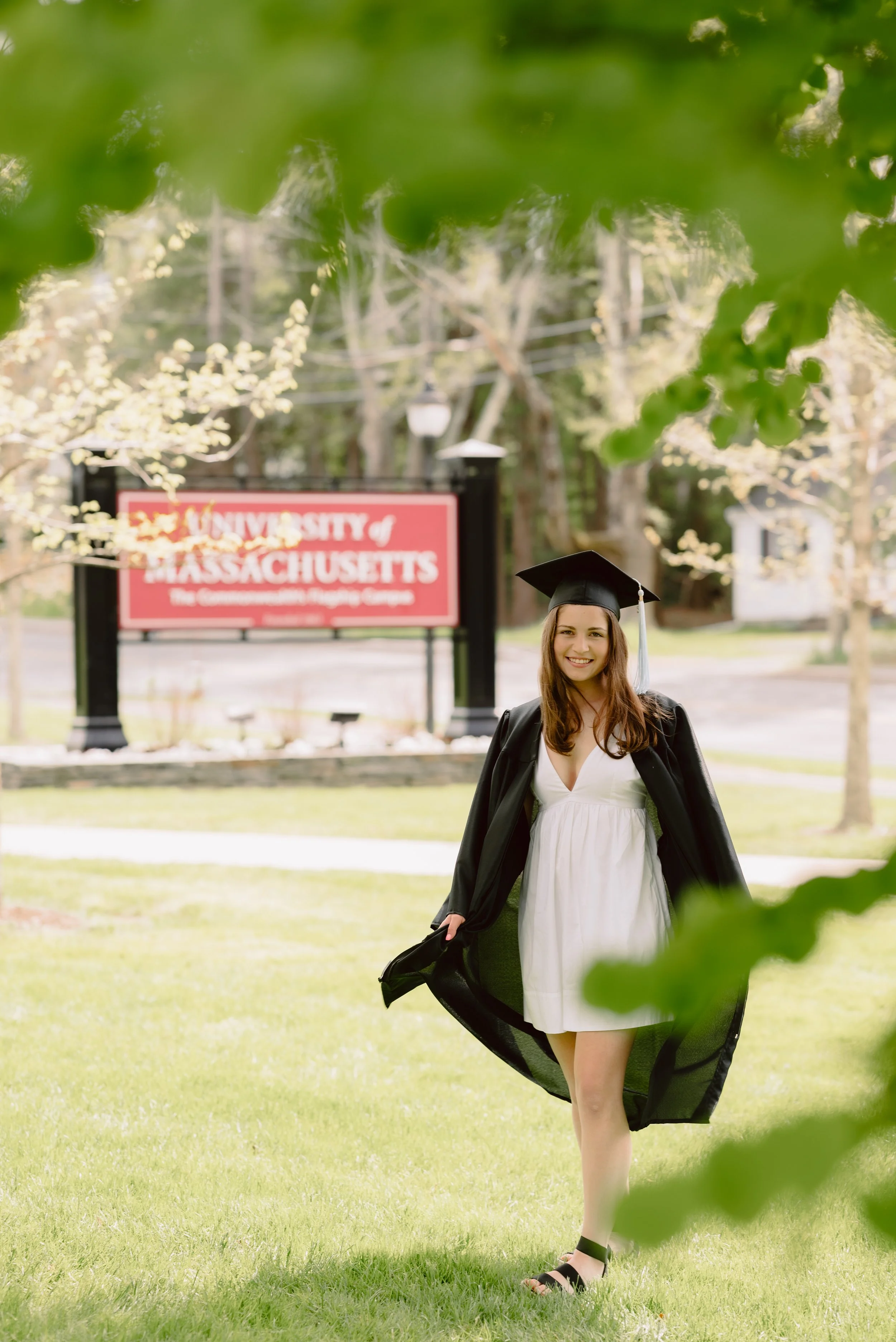 A young woman in a white dress and black graduation gown and cap standing outside on the campus of the University of Massachusetts, smiling with a university sign in the background.