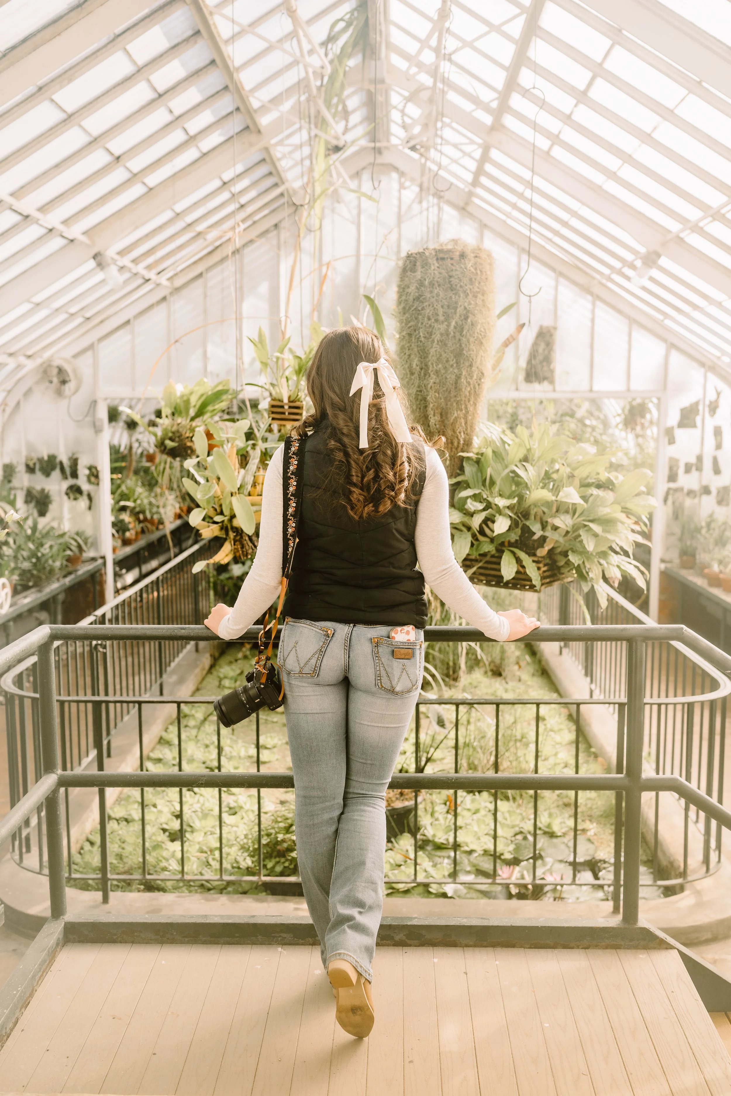 A woman standing inside a greenhouse, facing away, observing plants and flowers with sunlight streaming in.