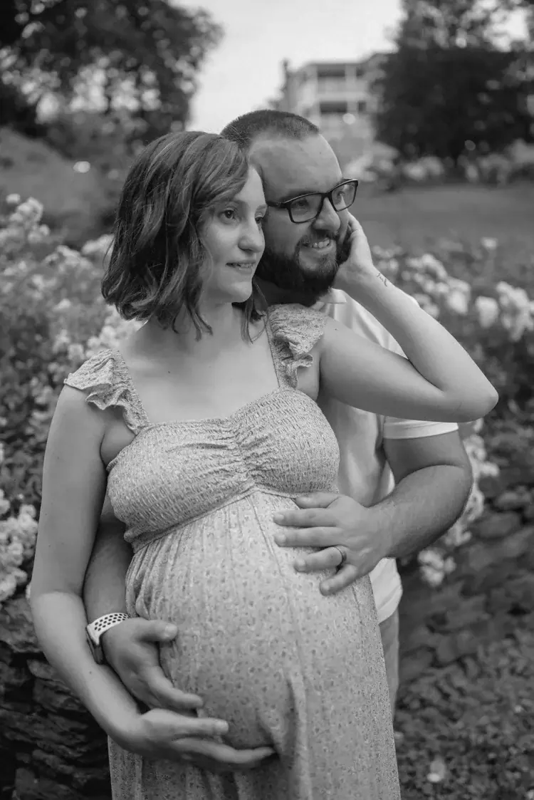 A pregnant woman and a man stand outdoors in a garden, with the man resting his head on her shoulder and both smiling. The woman is wearing a light-colored dress and the man is wearing a white shirt and glasses.