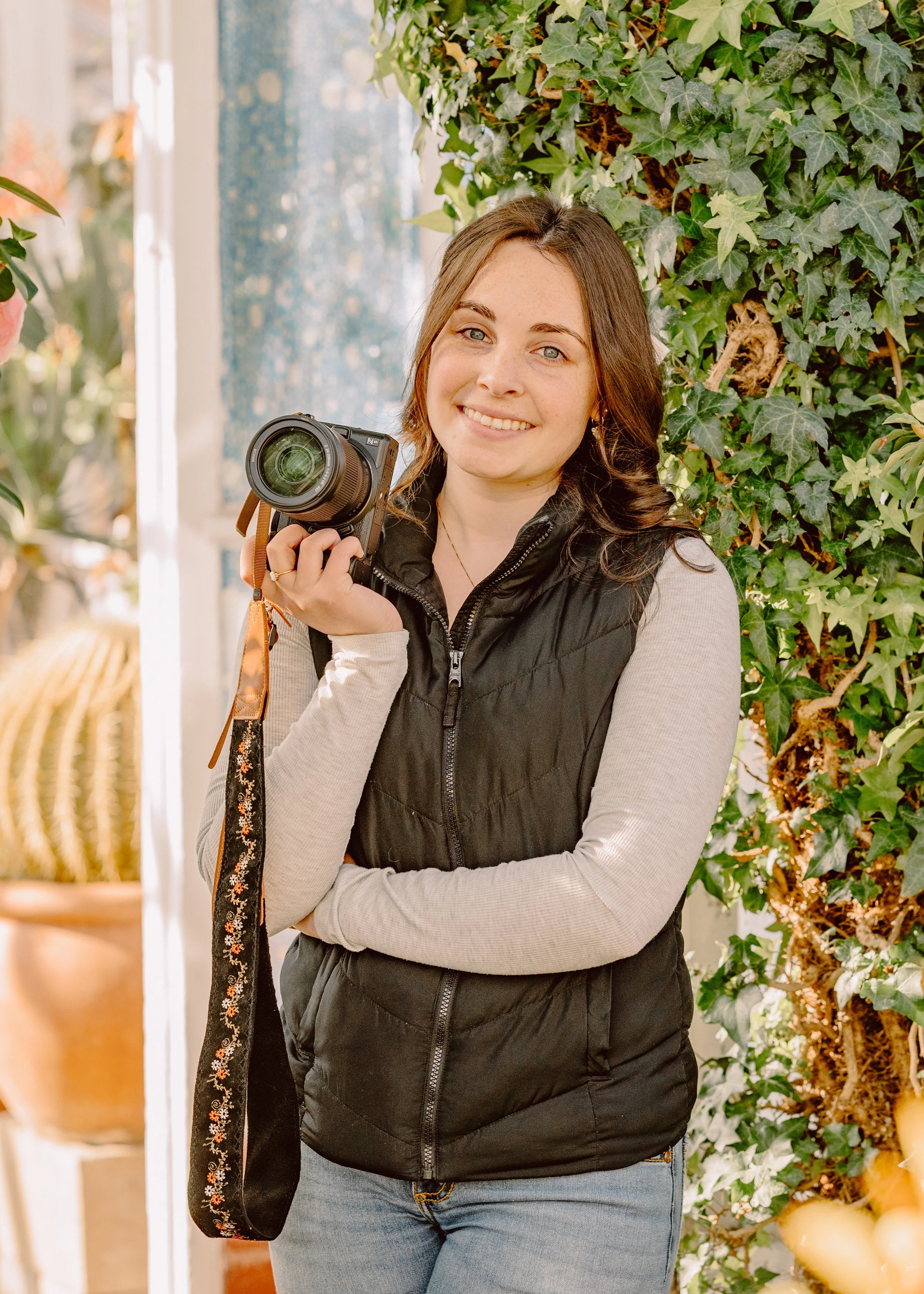 A young woman with brown hair, smiling, holding a camera in her right hand, standing against a background of green ivy plants.