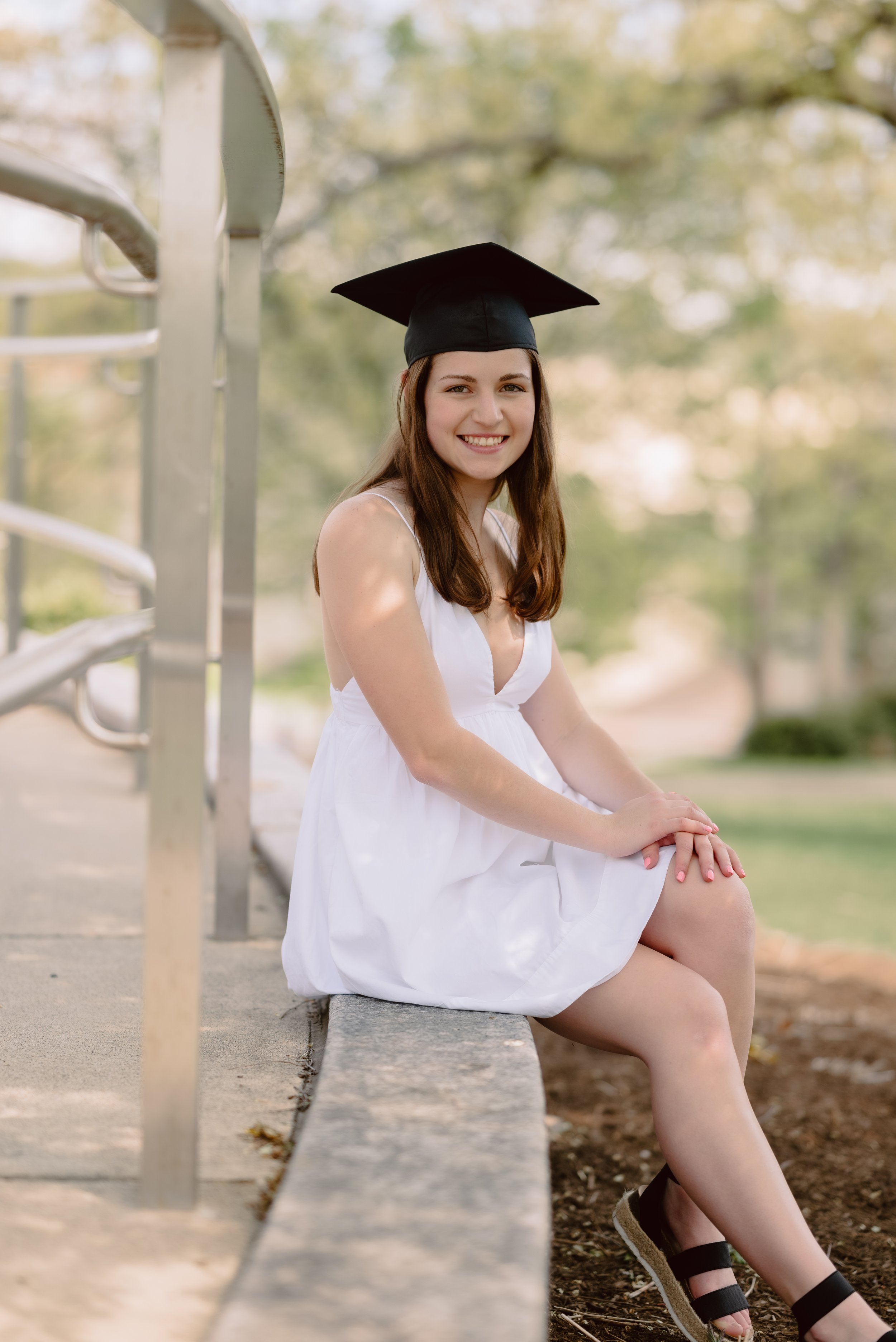 A young woman in a white dress and a graduation cap sitting on a concrete bench outdoors with trees in the background, smiling at the camera.