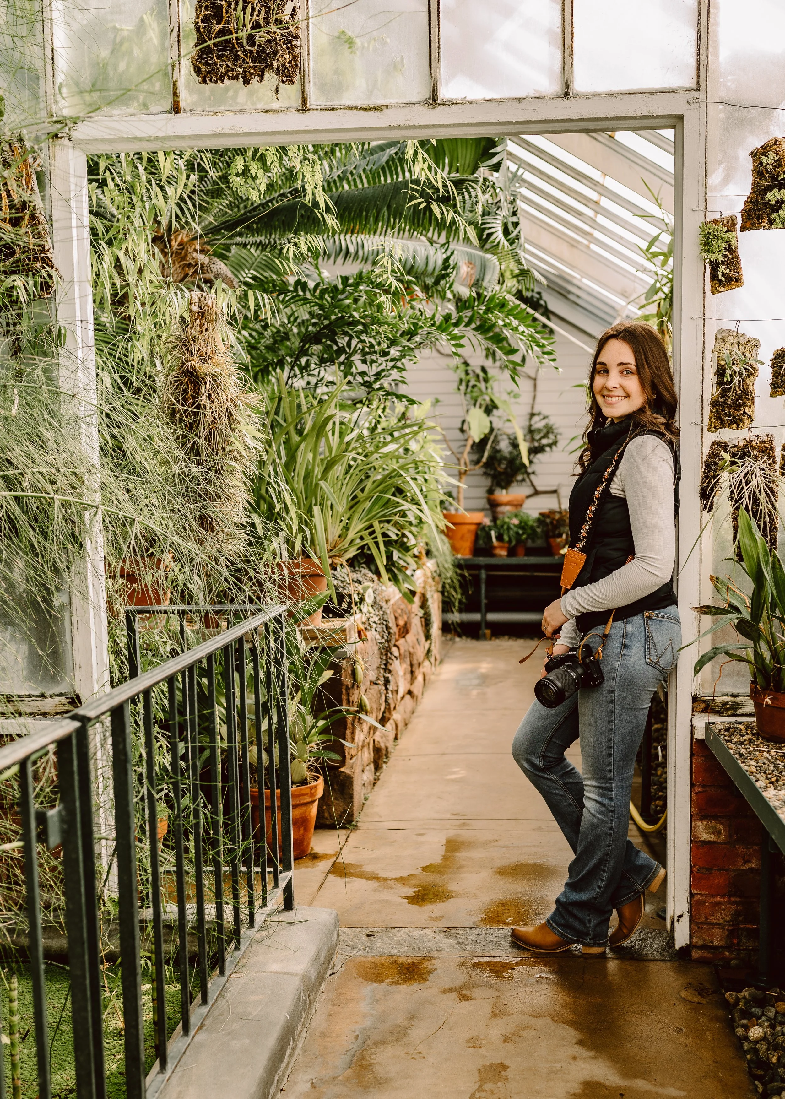 A smiling woman holding a camera stands in a greenhouse surrounded by various potted plants and greenery.