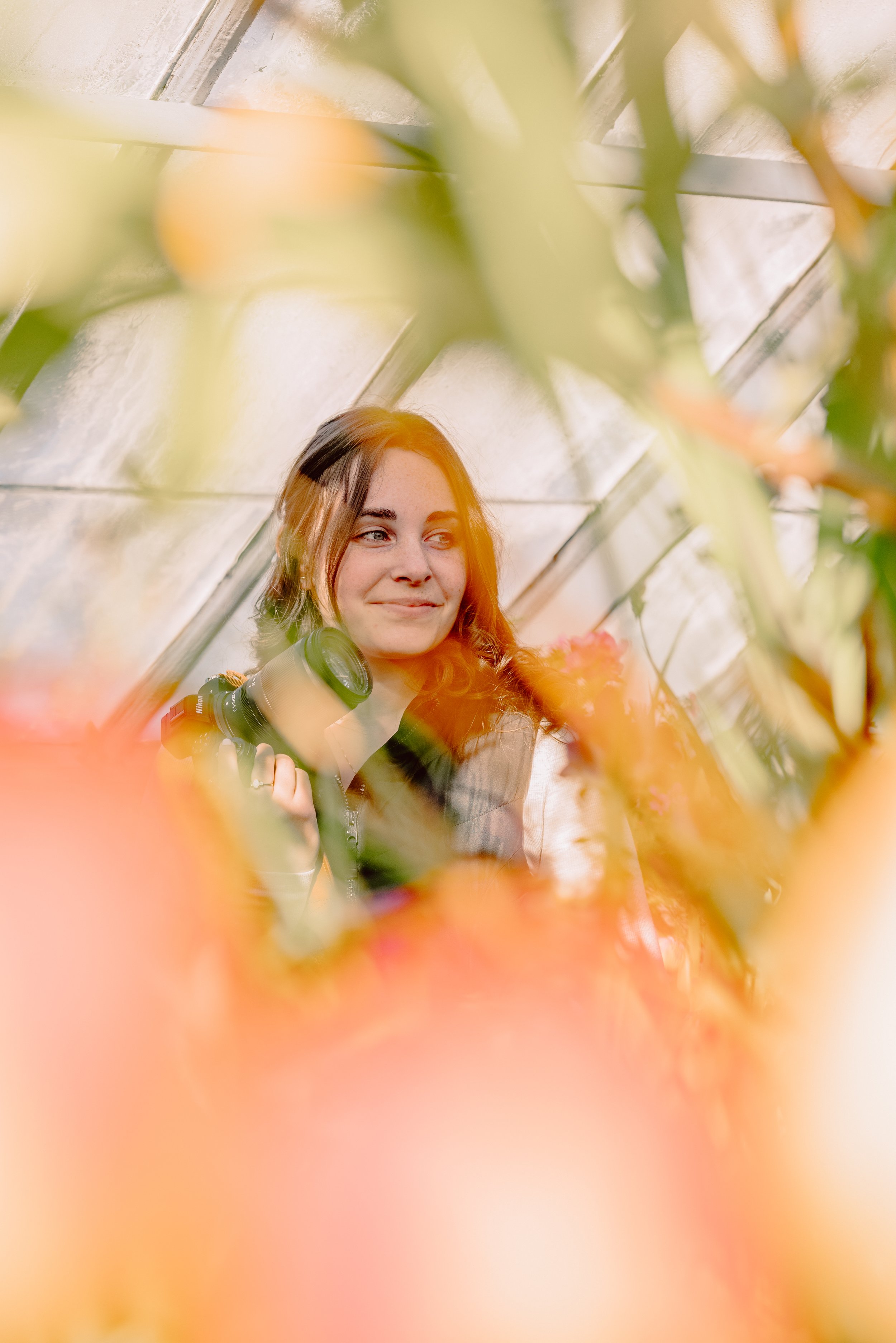 A woman with light brown hair holding a camera is seen through orange flowers in a greenhouse or garden.