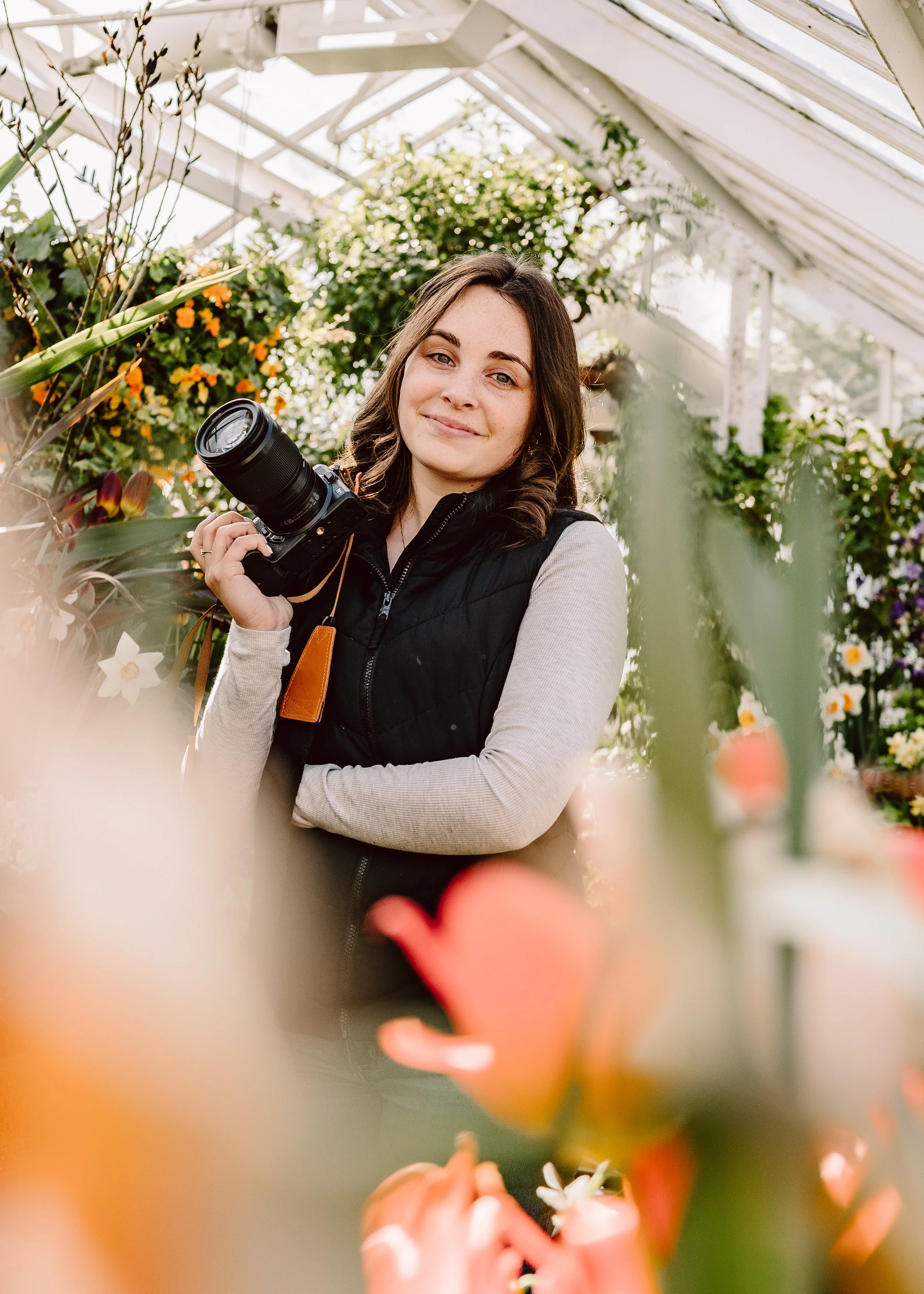 A woman with brown hair holding a camera inside a greenhouse filled with colorful flowers and green plants.