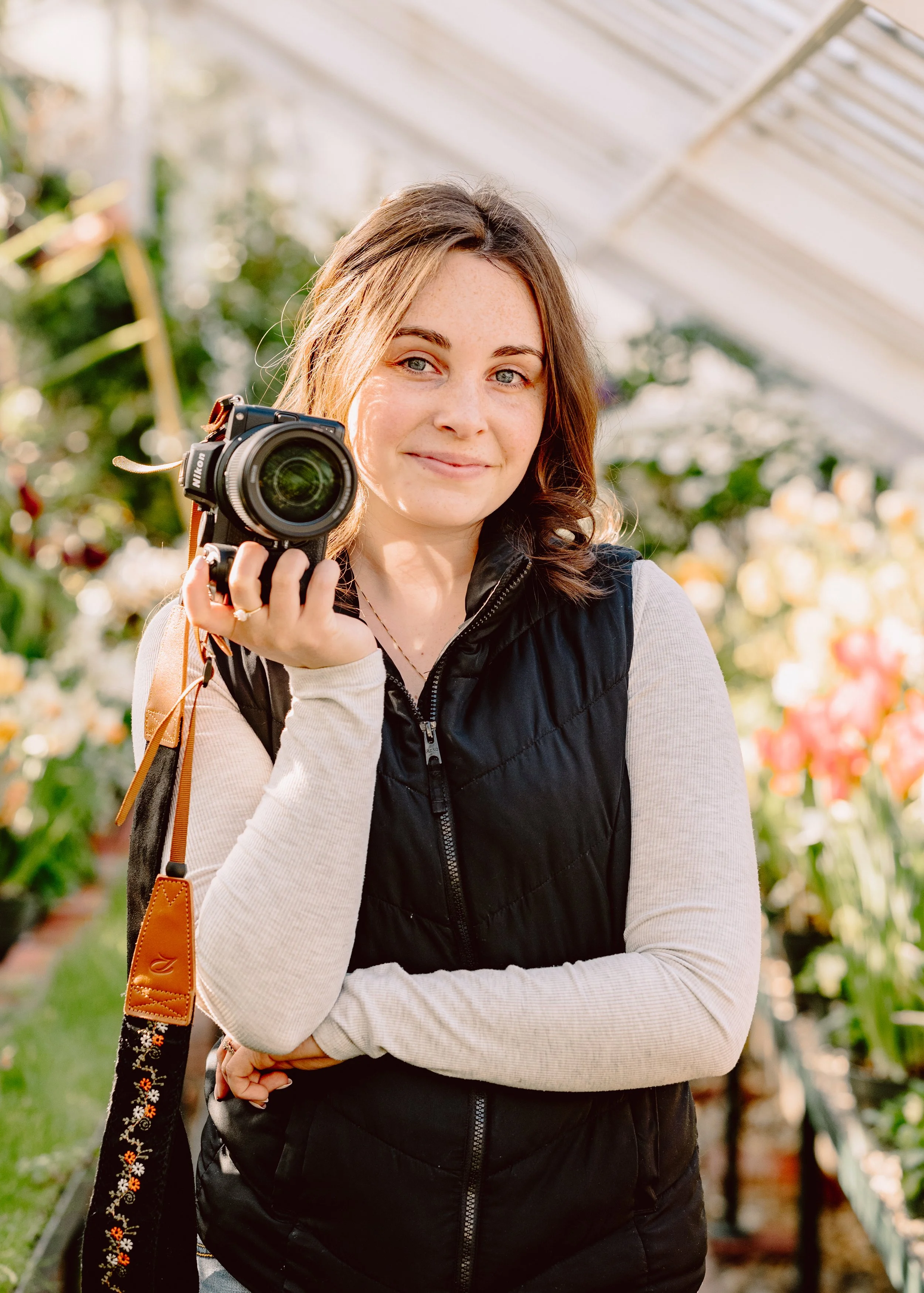 A woman stands in a greenhouse holding a camera, smiling, with flowers and greenery in the background.