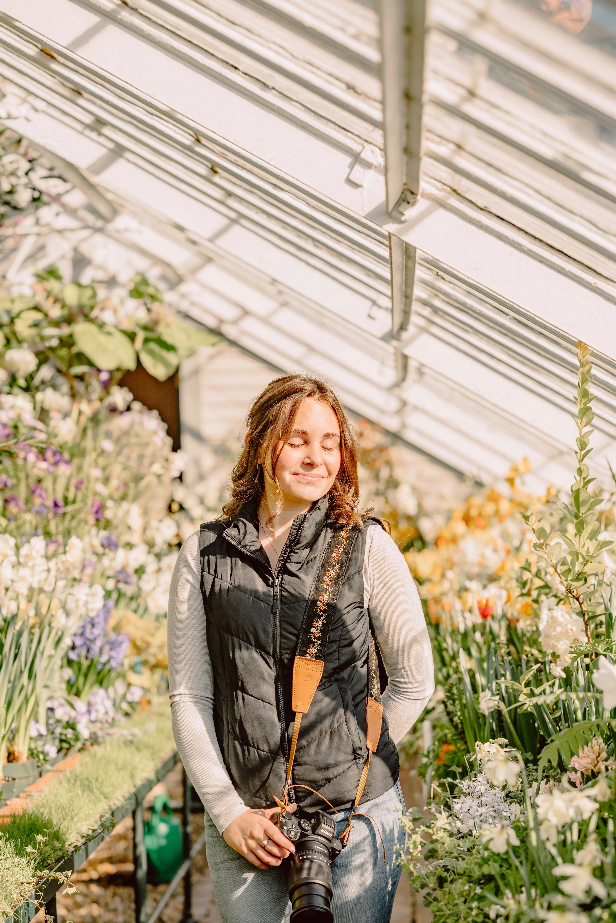 A woman with brown hair wearing a black vest and gray long-sleeve shirt holds a camera while standing in a greenhouse surrounded by blooming flowers.