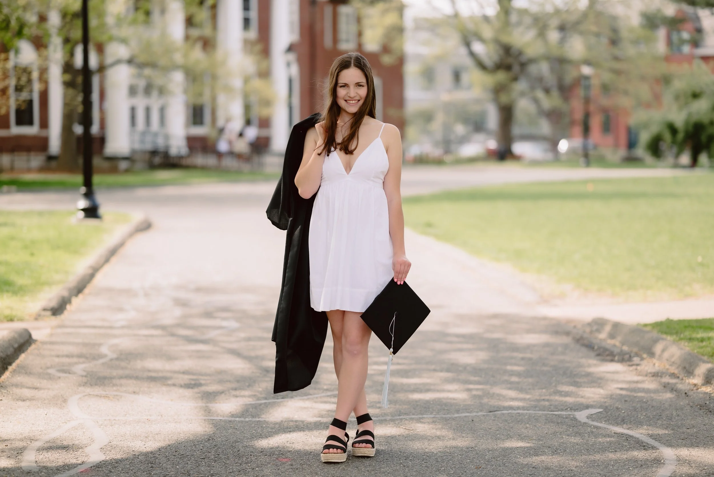 A young woman in a white dress holding a graduation cap, standing on a campus sidewalk, with buildings and trees in the background.