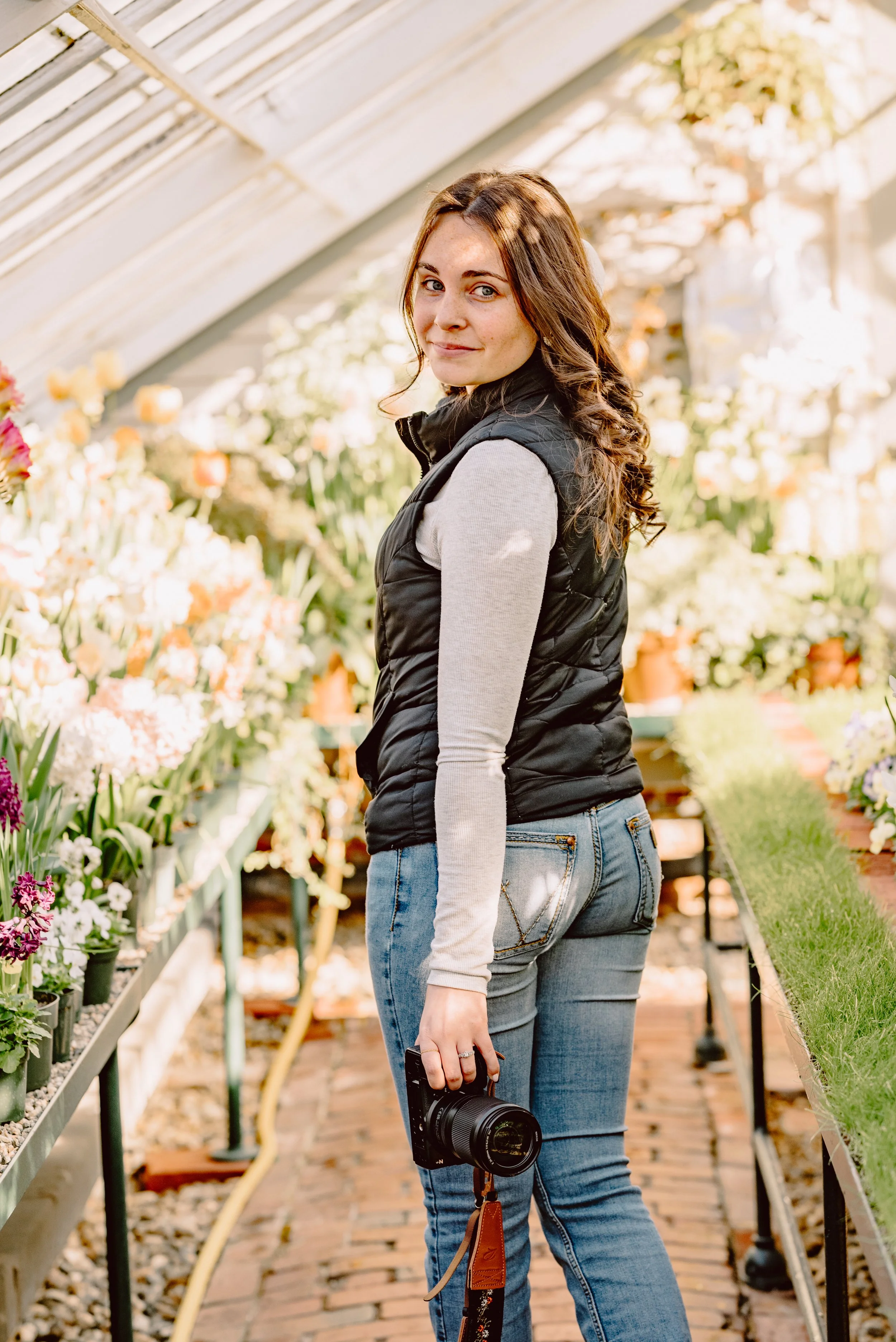 A woman standing in a greenhouse surrounded by flowering plants, holding a camera in her right hand.
