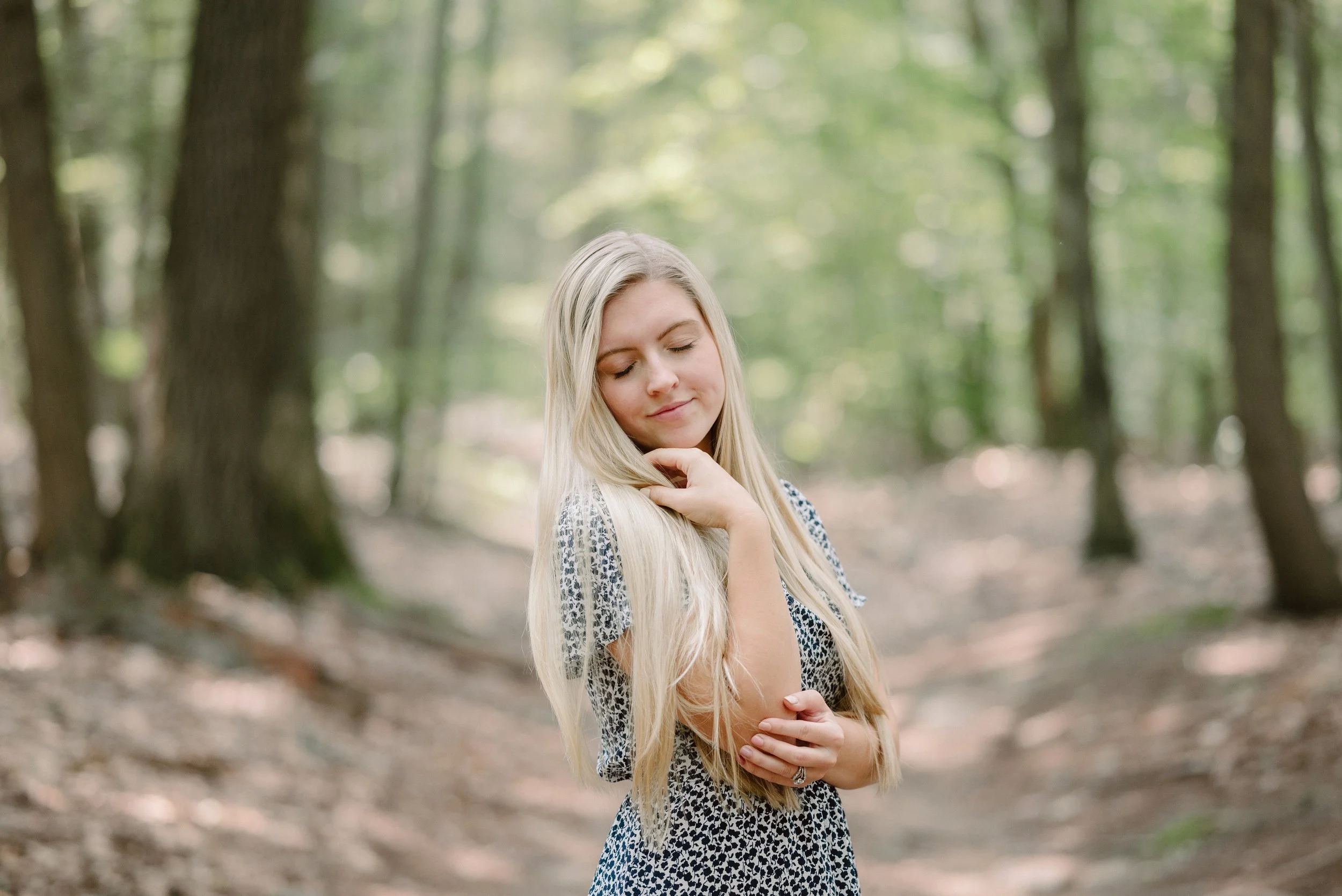 A young woman with long blonde hair standing in a green forest, smiling softly with her eyes closed, wearing a patterned dress.