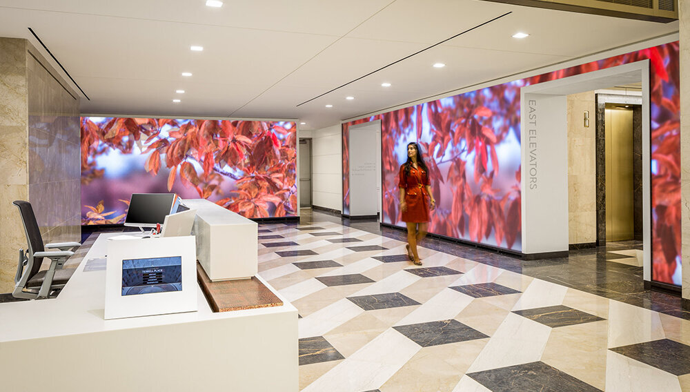 A woman walks through the bright lobby of an office building with large walls of moving videos of plants swaying.