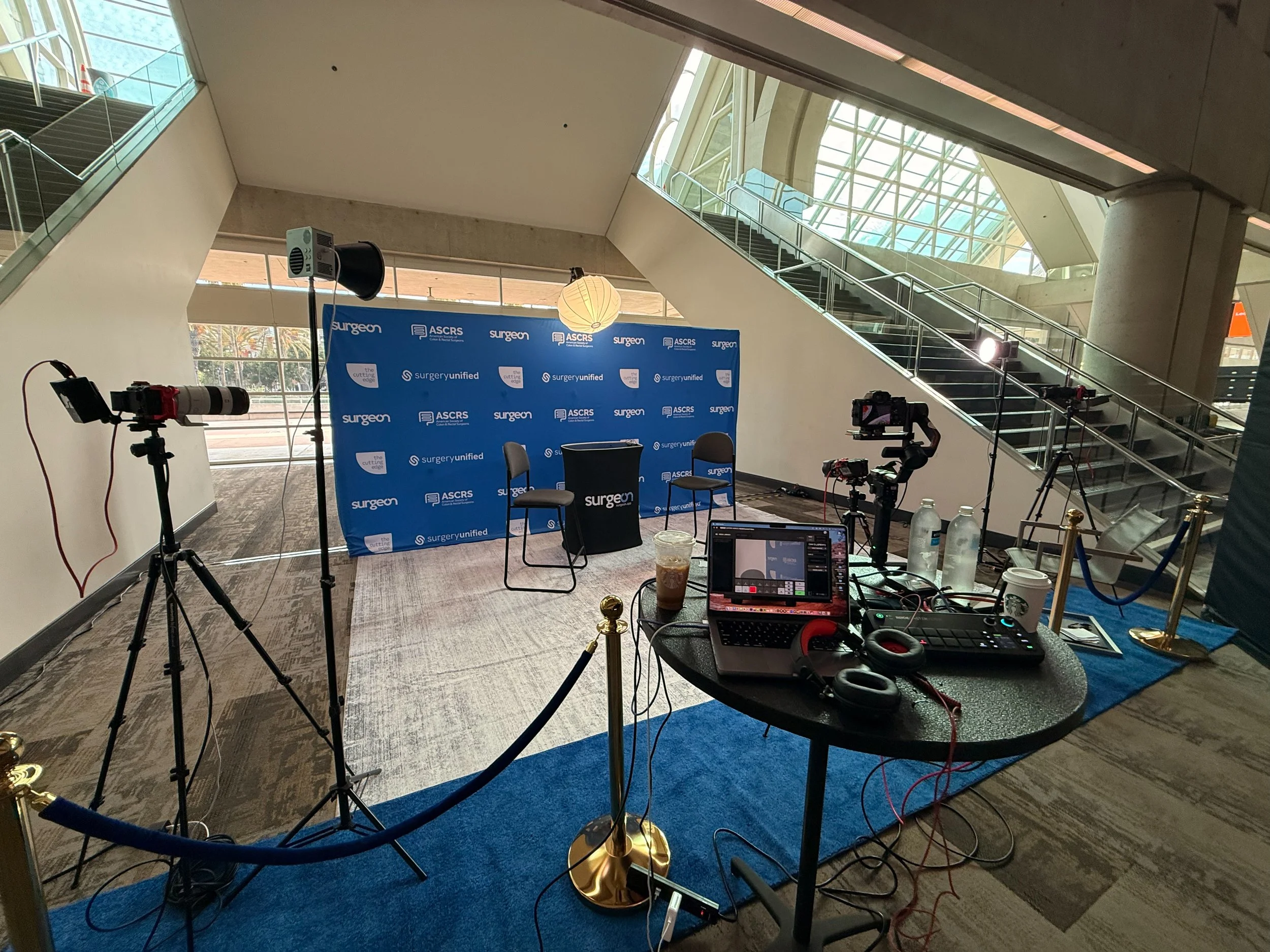 Setup for a virtual event or interview with a backdrop displaying 'surgery' and 'ASCRS' logos, including cameras, lighting, and a laptop on a table for recording or streaming.